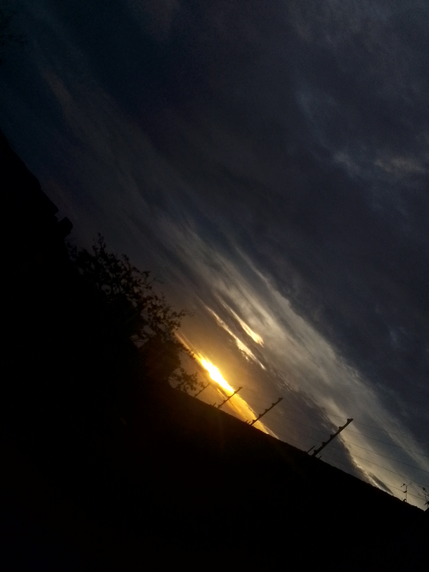 The photograph depicts a sunset in Juan Griego, Venezuela. The sky is mostly dark with visible clouds. A bright yellow sun is near the horizon, partially obscured by a dark land mass. Silhouettes of trees and what appear to be utility poles can be seen along the horizon. The image is taken at an angle, which suggests the photographer was standing on a slope or hill.