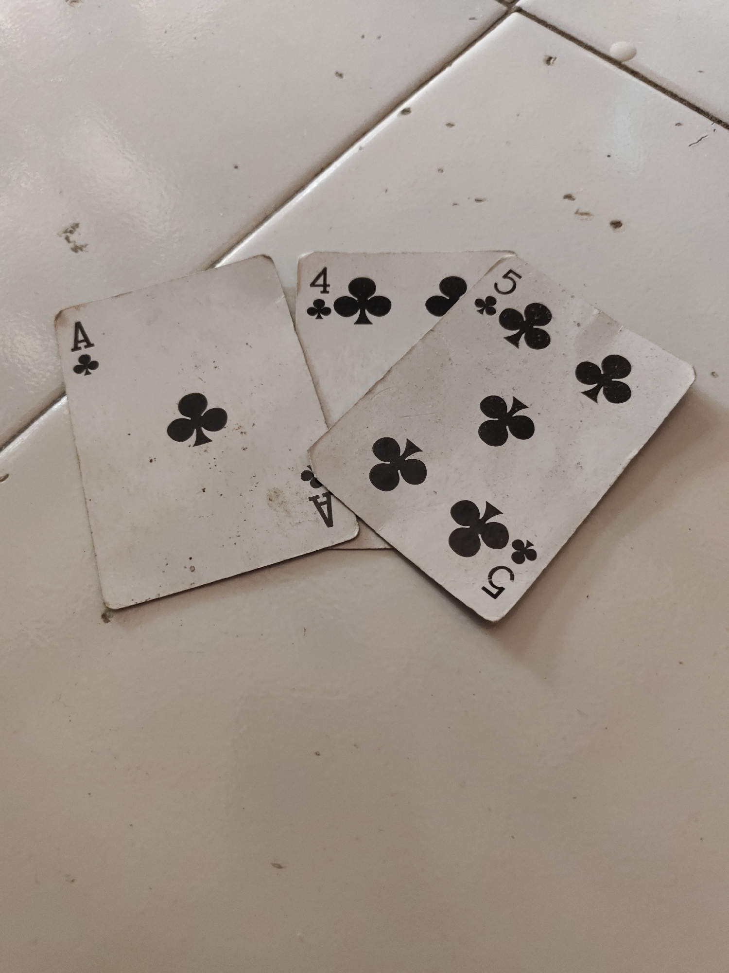 Three playing cards are scattered on a tiled surface. The cards are all of the club suit. One card is an Ace of Clubs, another is a 4 of Clubs, and the third is a 5 of Clubs. The cards appear worn and have some dirt or smudges on them. The tiled surface is light-colored and appears to be outdoors, possibly on a patio or sidewalk. The image is taken from a slightly elevated angle, looking down at the cards. There is no indication of the specific location in Bangsalsari, Indonesia, other than it being an outdoor tiled surface.