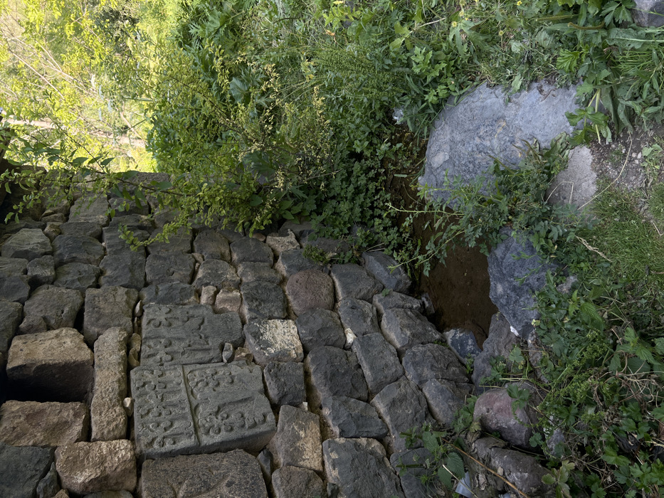 A stone wall with embedded engraved blocks is partially covered with green plants. The stones are varying shades of gray. There are trees and foliage at the top of the image. A large gray rock formation with a dark hole is to the right. Plants grow on and around the stones and rocks.