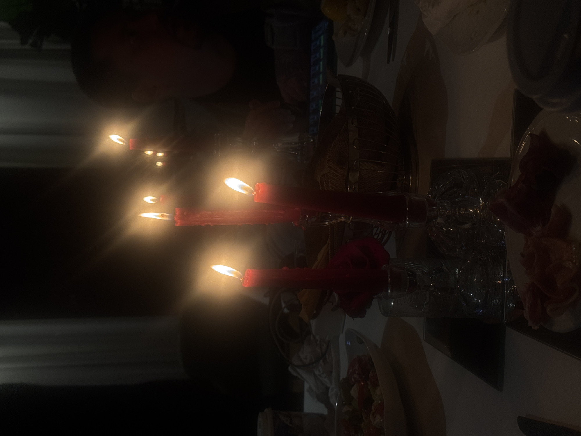 The image depicts a dimly lit dining table setting, likely indoors, with several red candles burning in glass candle holders. A floral arrangement, potentially roses, is present near one of the candle holders. Plates with food, including what appears to be prosciutto, are visible on the table. A person's head is faintly visible in the background, but it's difficult to discern details. A metal basket or serving dish also sits on the table. The location in Moscow, Russia cannot be verified from the visual data provided.