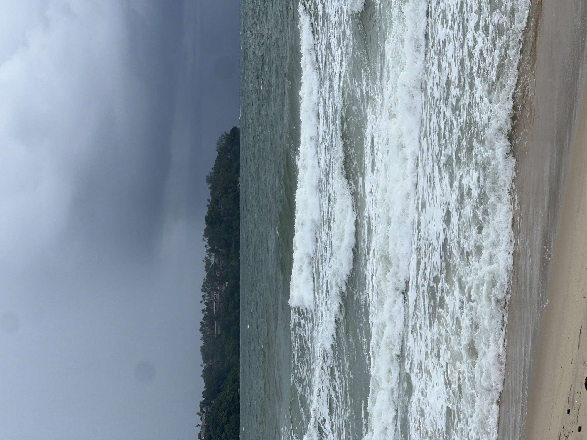 The media file displays a coastal scene dominated by a powerful, turbulent ocean and an overcast sky. The image is oriented vertically, but depicts a horizontal landscape.
In the center, large, white-capped waves crash and roll towards a sandy beach. The ocean water appears a deep, somewhat muted green-grey, reflecting the heavy sky. To the right, a strip of light brown, wet sand forms the beach, where the waves are receding and breaking.
The upper portion of the scene is filled with a vast expanse of dark grey, heavy clouds, suggesting an overcast day, possibly indicative of an impending storm or just very cloudy weather. To the left, a distant landmass rises from the sea, densely covered with dark green trees. Several small structures, likely buildings or residences, are visible nestled within the foliage on this distant hillside.
There are no people or visible text in the image. The scene captures the raw energy of the ocean under a dramatic sky, evoking a sense of nature's power and a somewhat moody atmosphere.
