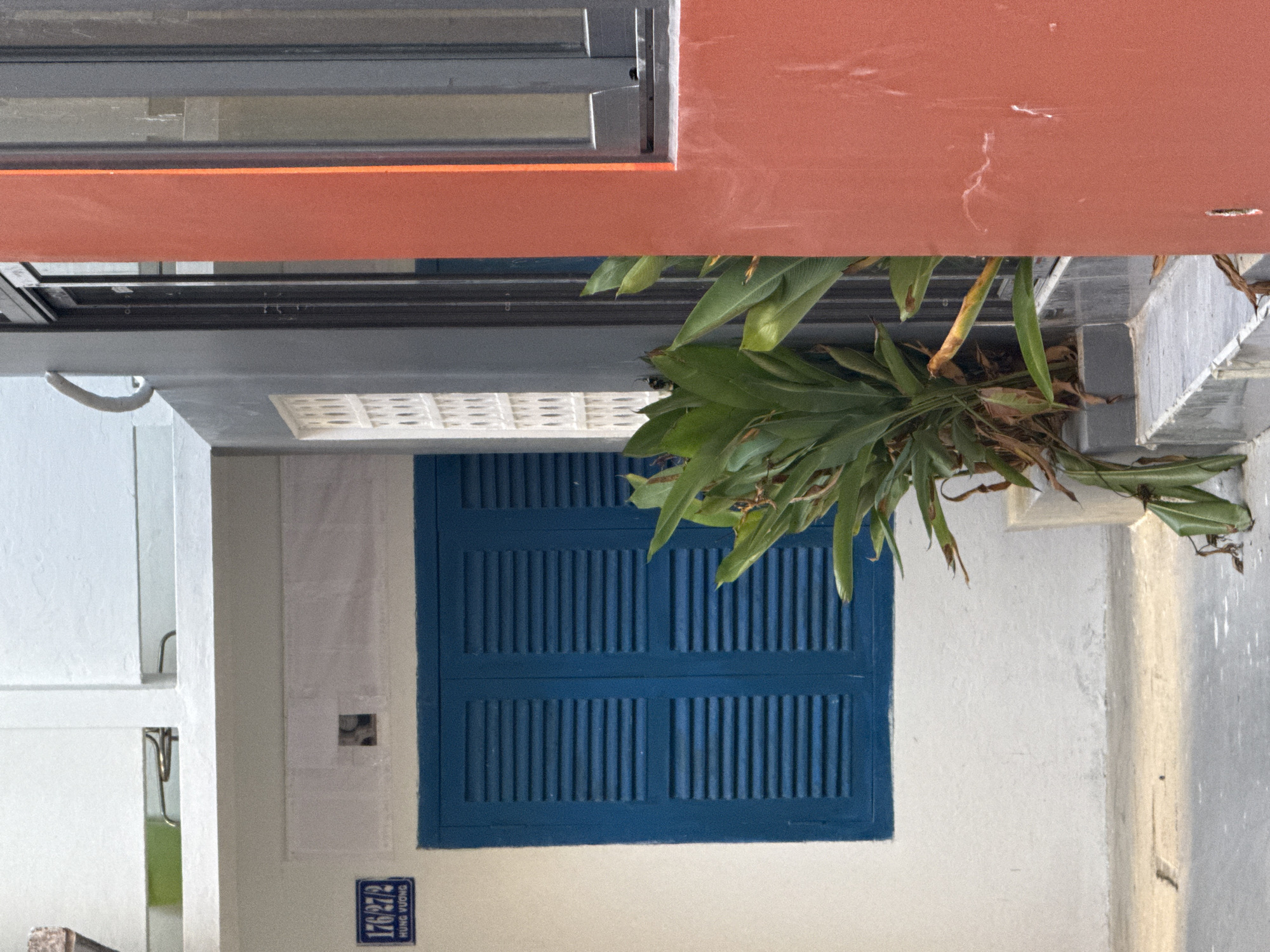 A building facade in Nha Trang Ward, Vietnam, is shown. A blue, shuttered window is centrally located on a white wall. A plant hangs above the window. A small street address plaque, with the number "176/27/2 HUNG VUONG," is positioned near the lower left corner of the window. Above, an orange horizontal architectural element is partially visible. The top of a window is visible above this element.