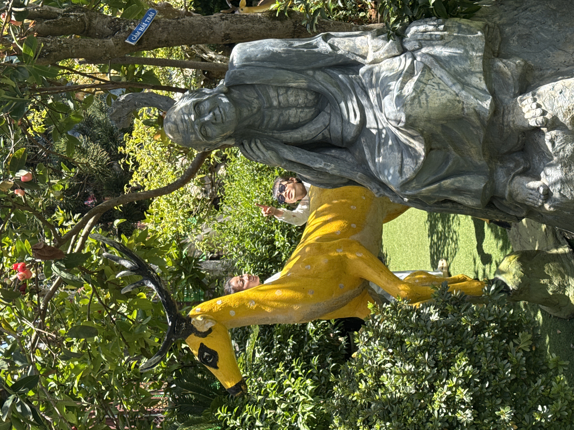 In Xuân Thành, Vietnam, a sunny outdoor scene features a stone statue of an elderly person seated in the foreground. To the left of the seated figure, a vibrant yellow deer statue with black antlers stands on a moss-covered rock. Behind the deer, two young individuals are visible. One, wearing sunglasses, points upwards with their left hand, while the other, with their face turned to the right, looks towards the deer. Lush green foliage surrounds the statues, and a tree with pink flowers grows in the background. A blue sign with white text is attached to the tree.