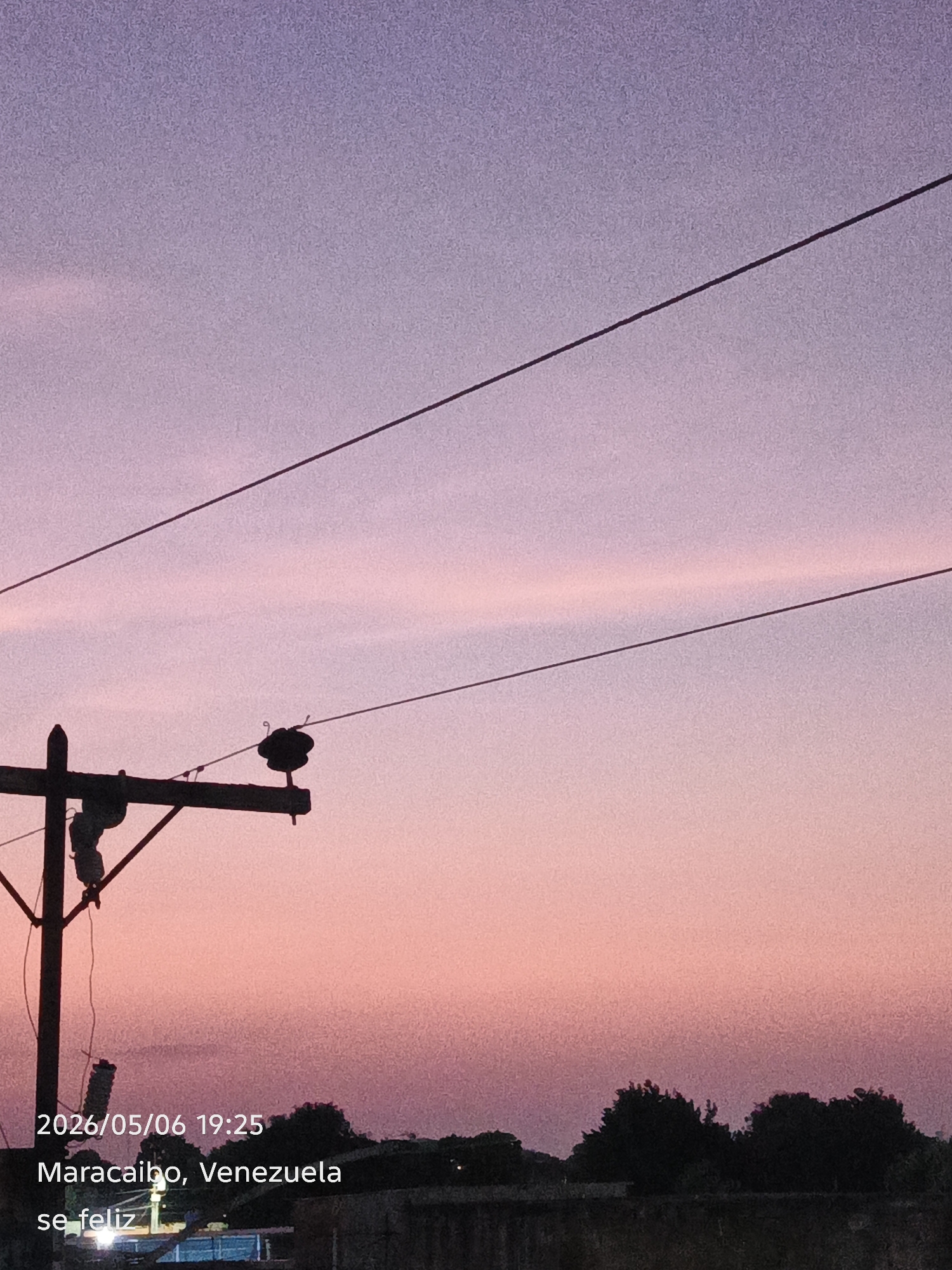The image shows a twilight sky transitioning from purple at the top to pink at the bottom. Two electrical wires stretch diagonally across the upper portion of the frame. A wooden utility pole is visible on the left side of the image. The lower part of the image shows silhouettes of trees and a low wall. Text at the bottom indicates the location as Maracaibo, Venezuela, and the date and time as 2026/05/06 19:25. A phrase "se feliz" is also present.