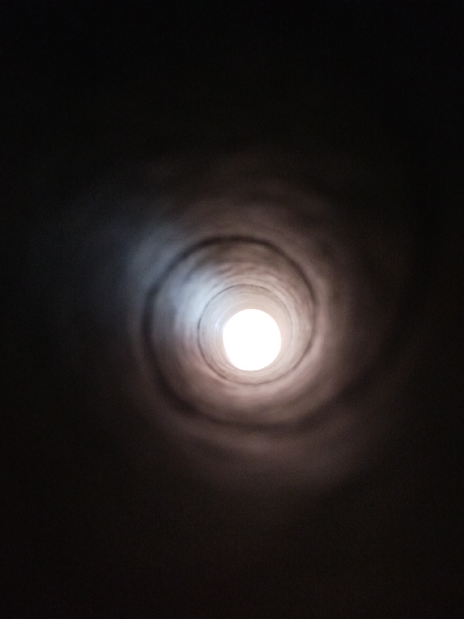 The image is a low-angle, indoor shot looking up into a dark, cylindrical space. The walls of the cylinder appear to be made of a textured material, possibly concrete or stone, and show evidence of wear or age. Concentric rings of light and shadow are visible, suggesting the curvature of the interior. At the center of the cylinder, a bright, white light source is visible, possibly an opening to the outside or a powerful artificial light. The overall impression is one of depth and enclosure. There is no indication of the specific location in Kremenchuk, Ukraine, in the image itself. The scene is devoid of people, objects, or any discernible action.