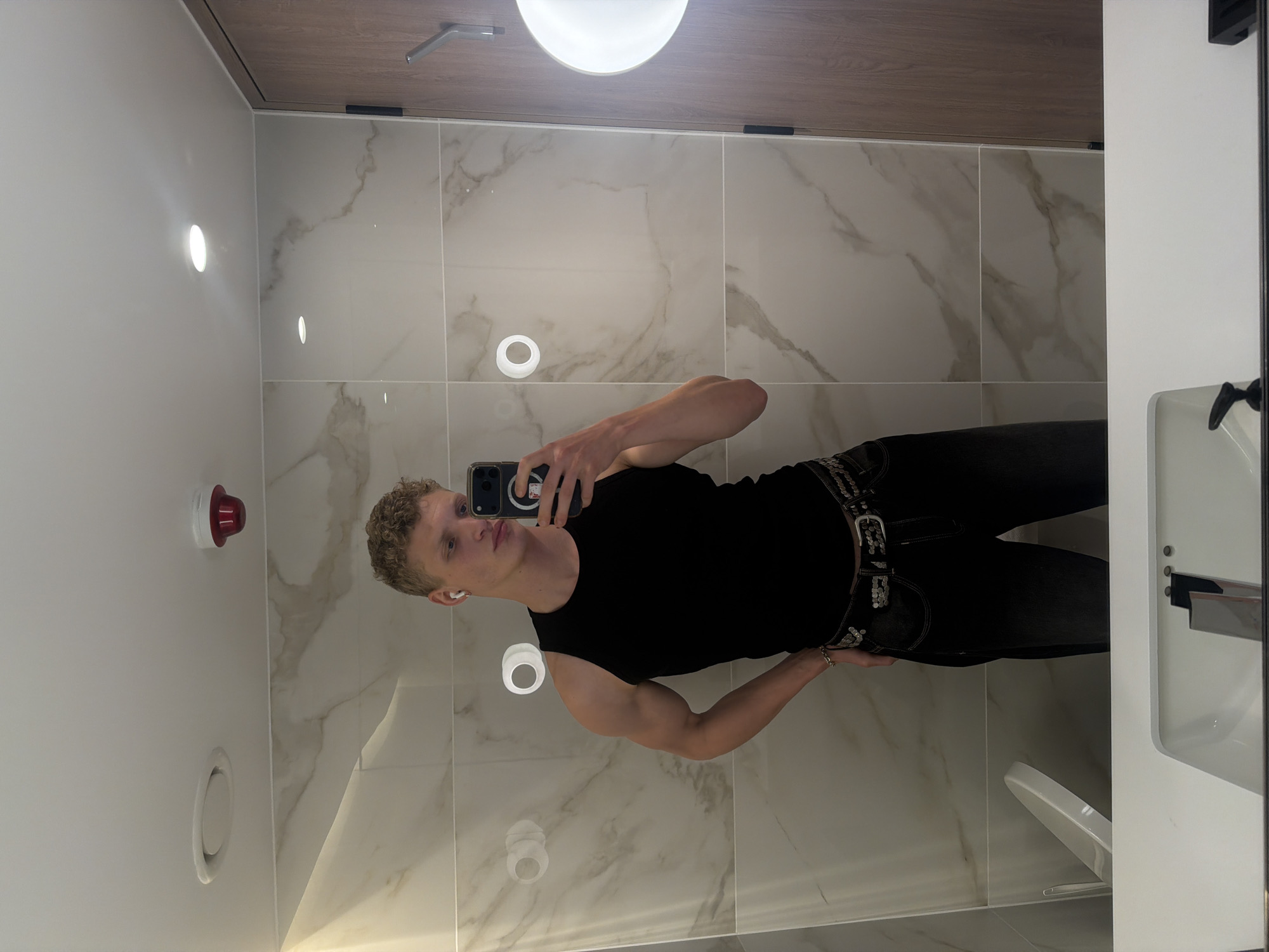 A man with short blond curly hair, fair skin, and wearing a black sleeveless shirt and black jeans with a studded belt is taking a selfie in a bathroom. He is holding a black smartphone in his right hand, and his left hand is on his hip. He is wearing white wireless earbuds in his left ear.
The bathroom has white walls with white marble-like tiles and a wooden ceiling. There are several circular lights on the wall and ceiling, and a red fire alarm is also visible on the wall. A white sink and a mirror are partially visible on the right side of the image.
The image does not provide sufficient information to pinpoint the exact location in the city of London, United Kingdom.