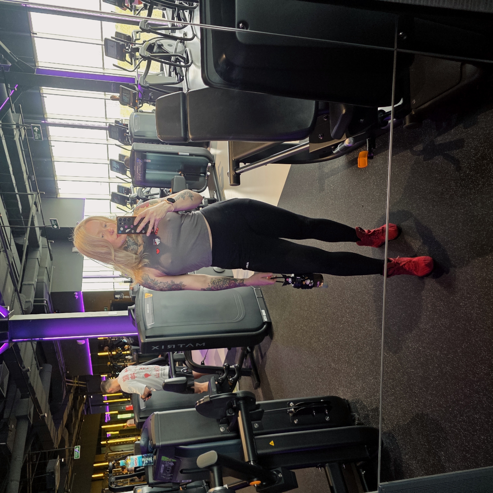 Here is a detailed description of the image:
The image captures a woman taking a selfie in a gym. She's holding her phone in her right hand, positioned to capture herself in a reflective surface, likely a mirror or a glass panel. She has light blonde hair, tattoos on her arms and neck, and is wearing a gray top, black leggings, and red sneakers. She is also holding a water bottle.
The gym setting includes various exercise machines such as ellipticals and weight machines. The lighting consists of both natural light from large windows and artificial lighting, with purple accents near the ceiling and warmer tones in the background. Another person is visible on a machine in the background wearing a white shirt. The floor is dark and the gym equipment appears modern.