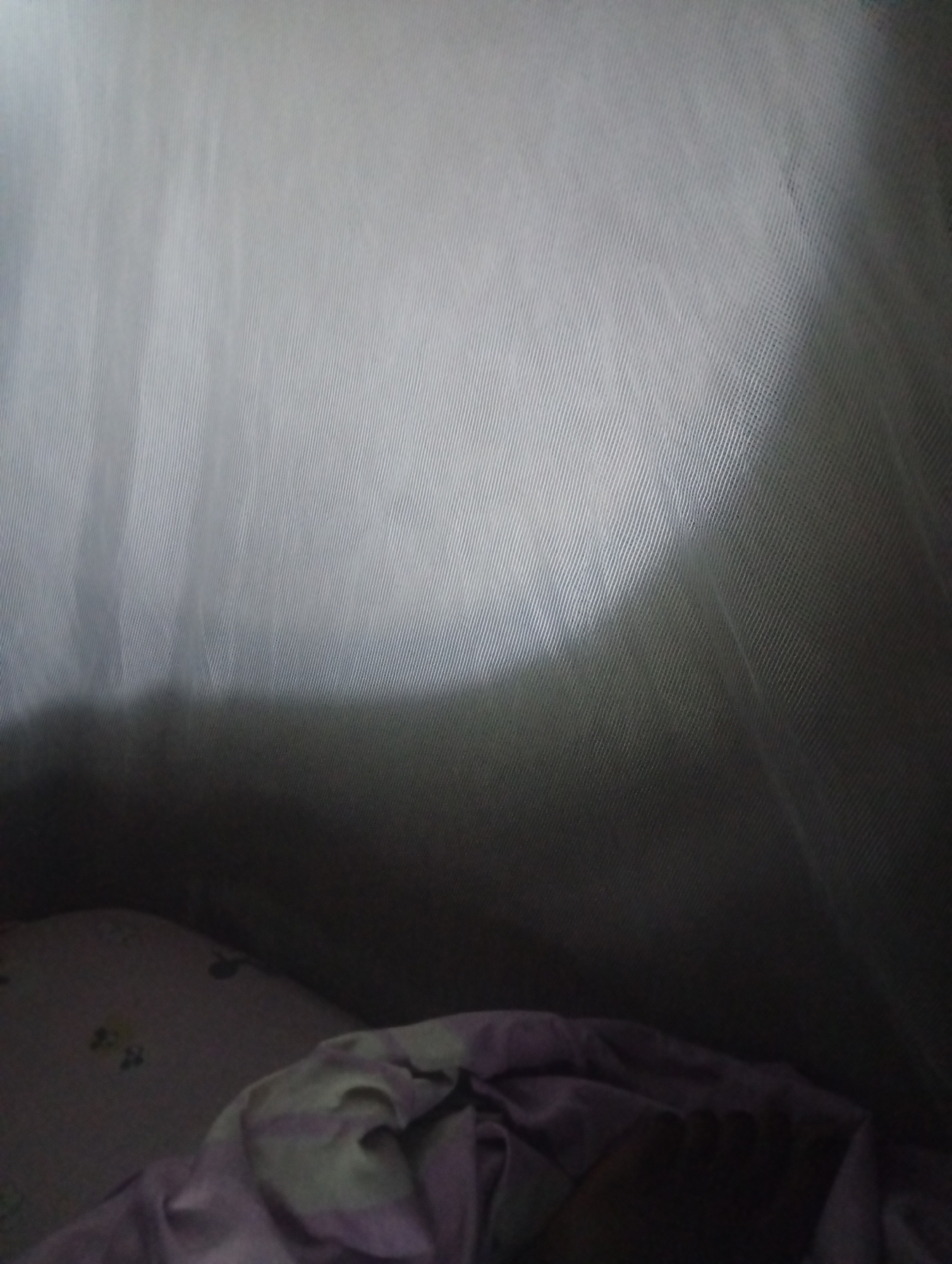 The image shows the interior of a tent. The tent fabric is light grey and has a ribbed texture. A shadow is cast across the fabric from the left. In the foreground, a pillow with a faint floral pattern and a purple and white striped blanket are visible. The scene appears to be inside a tent, possibly in a campsite. There are no people or actions depicted. The location in Jalingo, Nigeria is not discernible from the image itself.