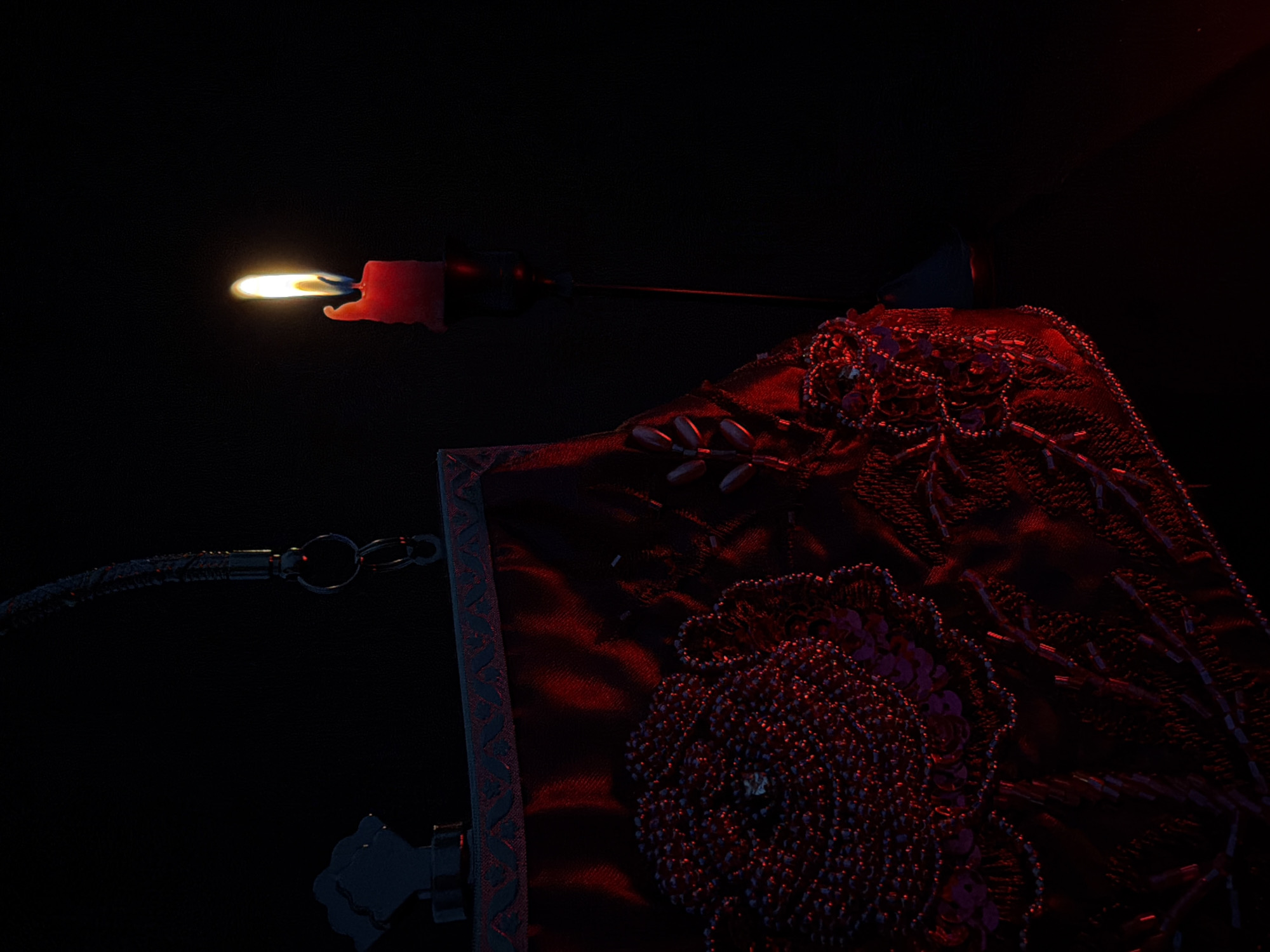 In a dimly lit environment with a black background, a red beaded clutch bag is positioned on the left side of the frame. The clutch is adorned with intricate floral patterns made of beads and sequins, with a metallic handle attached to its top. To the right of the clutch, a single red candle in a dark candlestick burns brightly, casting a warm glow. The candle flame is visible and illuminated against the dark backdrop. The scene appears to be an indoor setting, possibly a studio or a room, with no other visible elements or people. The overall mood is one of mystery and elegance, accentuated by the interplay of light and shadow.