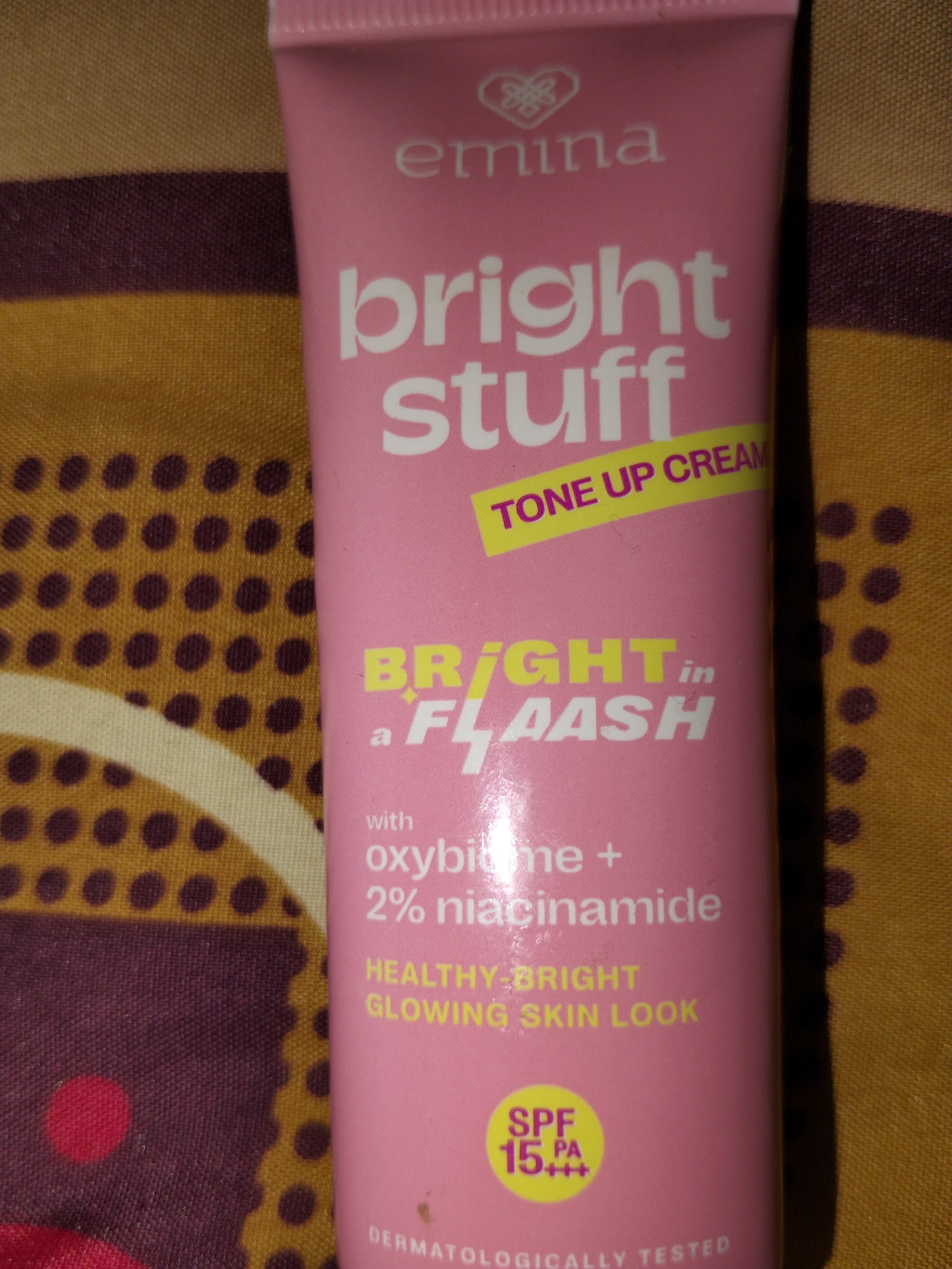 A close-up, eye-level, slightly angled shot shows the front of a pink tube of "Emina Bright Stuff Tone Up Cream." The tube has white lettering and a yellow band with the words "TONE UP CREAM" at an angle. Below this, larger white text reads "BRIGHT in a FLASH." Further down, smaller white text details "with oxybit me + 2% niacinamide" and "HEALTHY BRIGHT GLOWING SKIN LOOK." In the bottom right corner of the tube is a yellow circle containing "SPF 15 PA+++". The tube is positioned on a patterned fabric surface with alternating horizontal stripes of yellow and dark purple, and a pattern of dark purple dots on the yellow sections. The image does not contain any information about the city of Payagaboh, Indonesia.