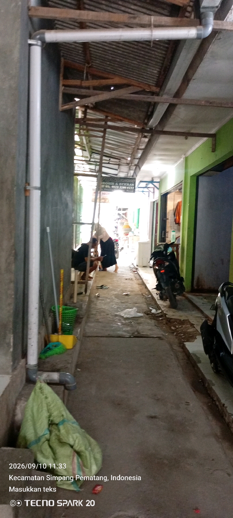 The image captures a narrow outdoor alleyway in Simpang Pematang, Indonesia. The scene is bordered by a grey wall on the left and a green building on the right. A corrugated metal roof, supported by wooden beams and metal brackets, covers a portion of the alleyway, providing shade.
In the foreground, a green sack lies crumpled on the ground. To its left, a white PVC pipe runs vertically along the wall, with a horizontal pipe branching off towards the roof. Nearby, a green plastic basket and a yellow dustpan are visible on a concrete ledge.
Further down the alley, two individuals are present. One person, wearing a black shirt and dark pants, is crouched down, looking at a mobile phone. Another person, wearing a light-colored top and a dark skirt, stands next to them, also appearing to be engaged with the phone. A motorcycle is parked behind them.
A sign with text is hanging from the roof structure in the middle ground, partially obscured by the angle. The ground surface is a mix of concrete and asphalt, with some scattered debris. The overall environment suggests a functional, possibly commercial or residential, back alley. The date stamp in the lower left corner indicates "2026/09/10 11:33".