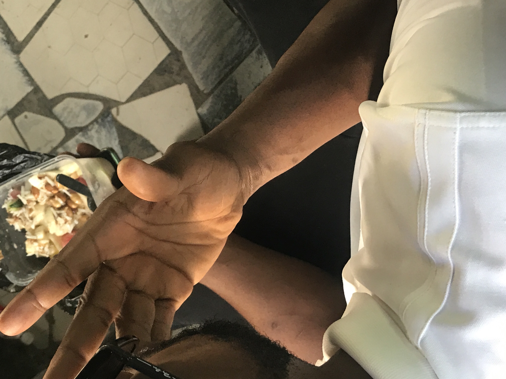 The image shows a close-up view of a person's arm and hand, holding a clear plastic container filled with food, possibly a salad or dessert. The person is wearing a white polo shirt. The background appears to be a tiled floor with a geometric pattern. The lighting suggests an indoor setting. There is no discernible information about the location in Kabong, Nigeria.