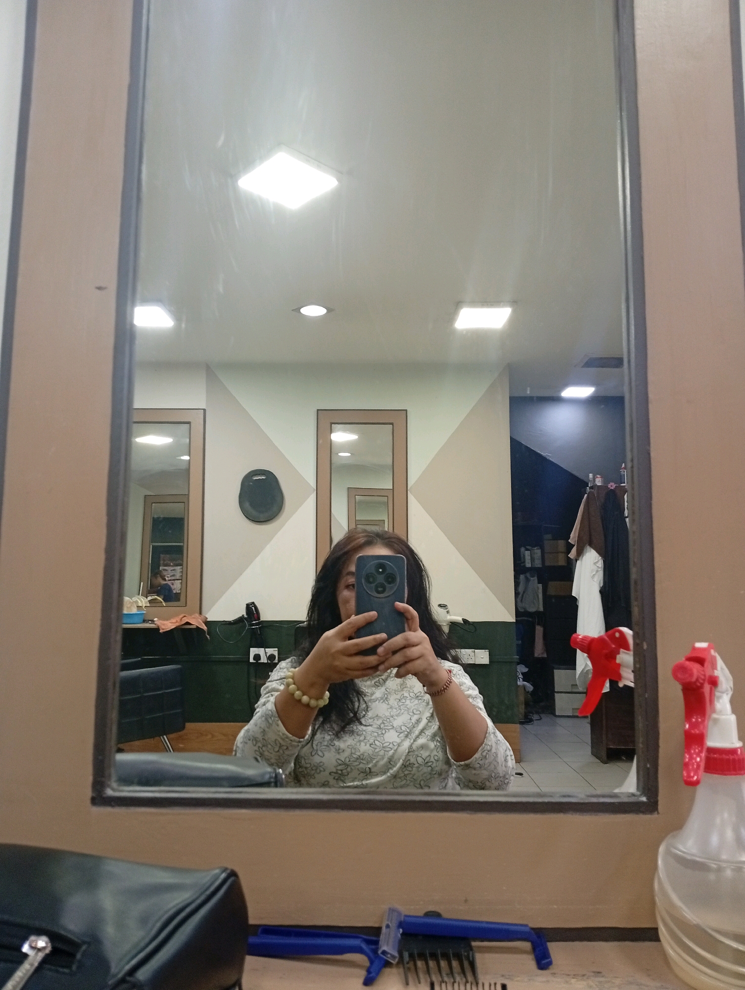 A mirror selfie is taken in what appears to be a salon in Selayang Municipal Council, Malaysia. The main subject is a person holding a smartphone and taking a picture of themselves in a large mirror. The person is wearing a patterned long-sleeved shirt and a bracelet. In the reflection, several salon chairs are visible, along with mirrors and a rack of clothing or towels in the background. The overall lighting suggests an indoor environment. On the counter in the foreground of the image, a black bag, a blue razor with a comb attachment, and two spray bottles (one red and white, the other clear with a red nozzle) are present.