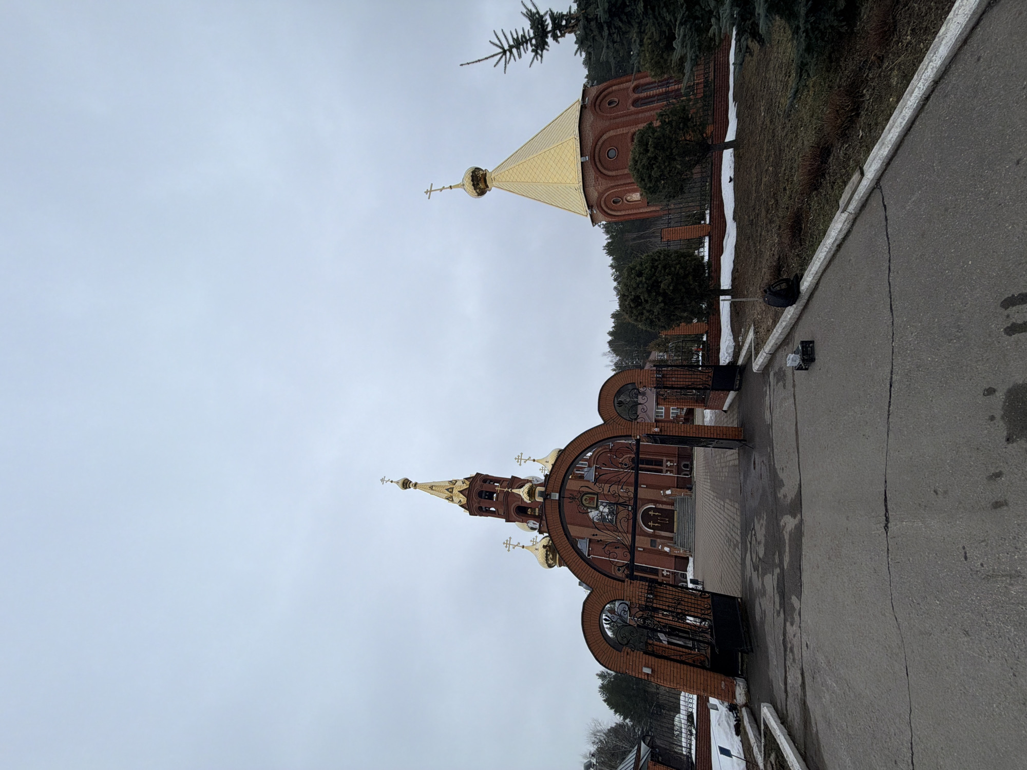 The image shows an external view of the Church of the Holy Great Martyr George the Victorious, located in the city of Chaikovsky, Perm Krai, Russia. The church is constructed of red brick, featuring multiple golden domes topped with crosses. A decorative wrought iron gate leads to the church entrance. A section of a pathway and a building with a golden roof can be seen beside the church. The sky appears overcast, suggesting a cold or cloudy day.