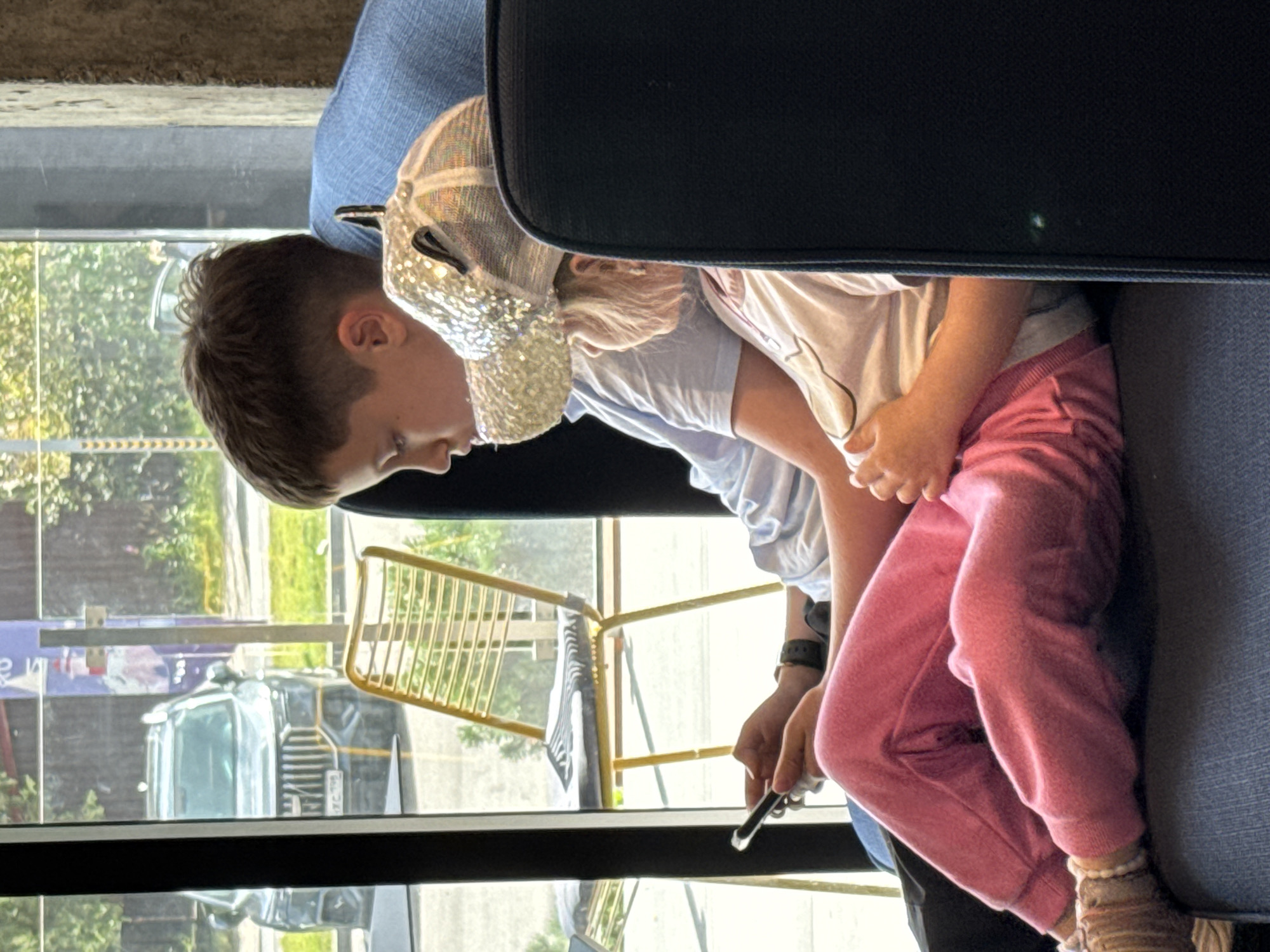 Two children are sitting in blue chairs, possibly in a cafe or waiting area. The older child, a boy, is on the left, looking down at a smartphone he is holding. He is wearing a light blue t-shirt and dark pants. The younger child, a girl, is to his right, wearing a light-colored t-shirt and pink sweatpants. She has a sparkly baseball cap on her head and is looking to the right. Through a window behind them, a gray SUV is visible parked on a street, along with a yellow chair and greenery. The overall scene suggests a casual indoor setting with a view of the outdoors. The location is identified as Lakhdenpokhya, Russia.