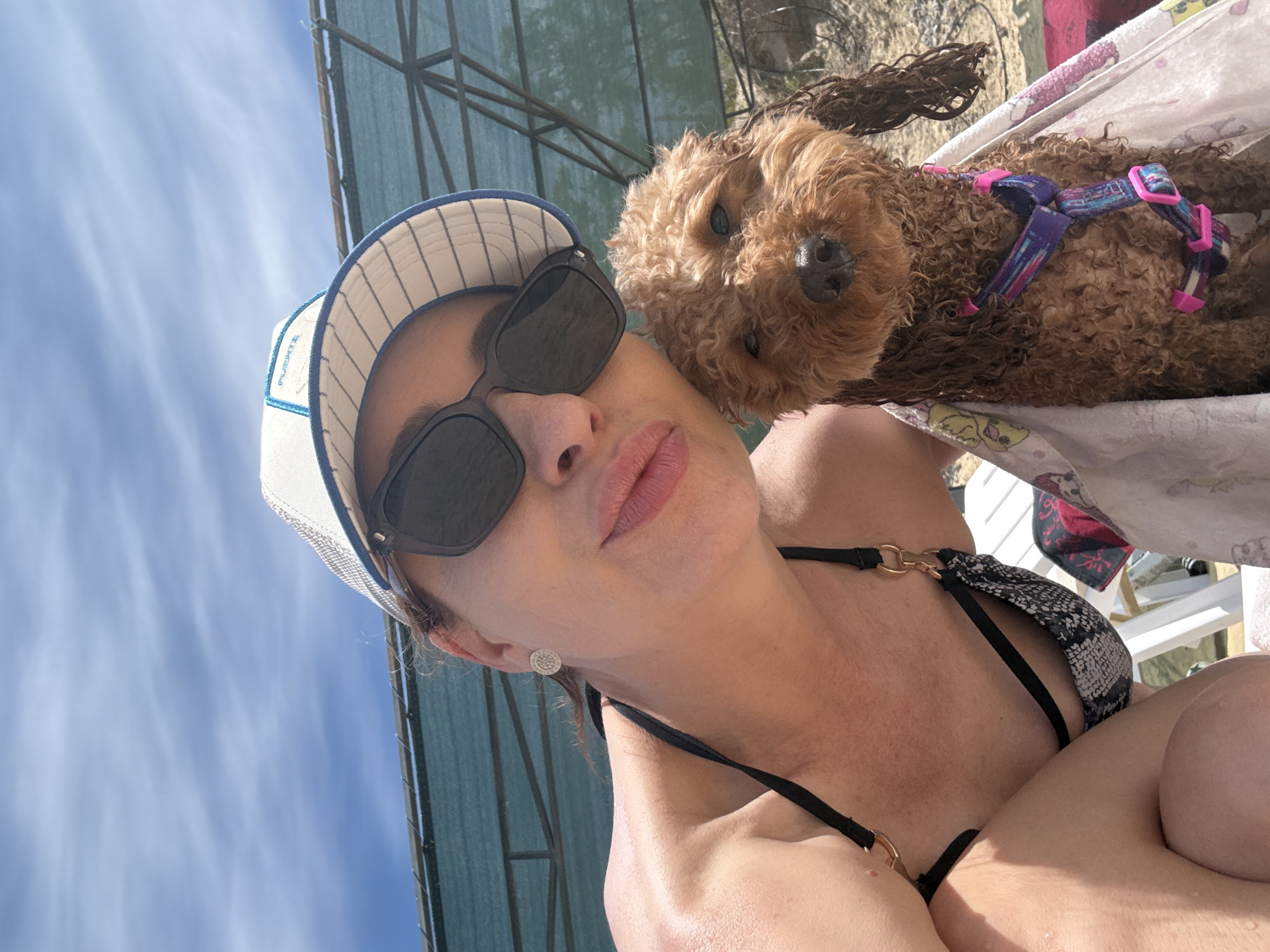 A woman with brown hair wearing a black bikini, a white and blue striped baseball cap, and dark sunglasses takes a selfie with a small brown poodle. The dog is wrapped in a white towel with cartoon characters and wears a colorful harness. They are outdoors, likely near a body of water, with a blue sky and clouds visible in the background, along with a green sunshade. The scene appears to be a leisure activity.