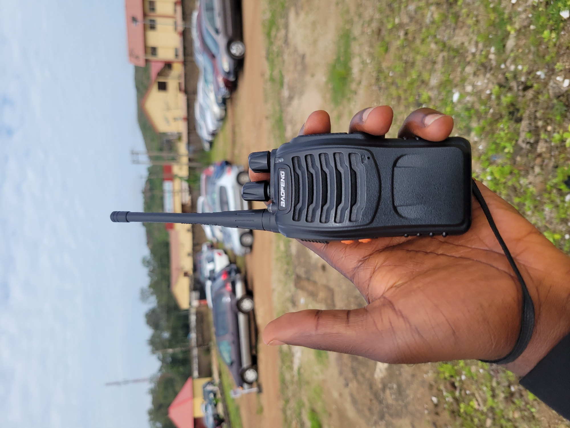 A close-up, eye-level shot shows a dark-skinned hand holding a black Baofeng walkie-talkie. The walkie-talkie's antenna is extended. The hand, with its thumb up, is positioned in the foreground, with the walkie-talkie in front of it. The background is an outdoor scene with a dirt lot, sparse grass, and a row of parked cars. Buildings with red and yellow roofs are visible in the distance, along with trees and a cloudy sky. The location is identified as Gura Top, Nigeria.
