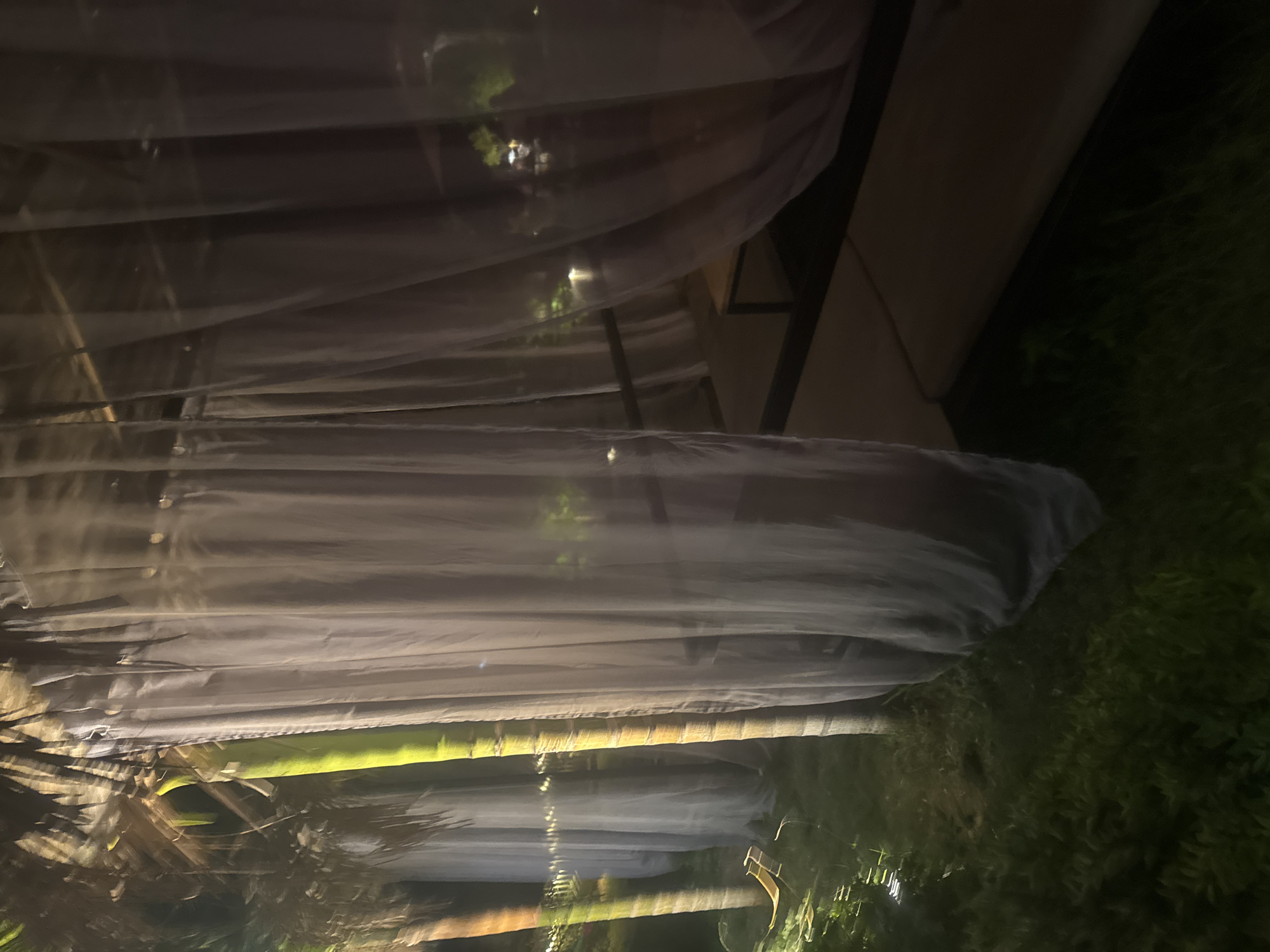 The image depicts an outdoor seating area at night, possibly a restaurant or resort in Bac Nha Trang Ward, Vietnam. The scene is characterized by rows of enclosed seating areas, draped with sheer white fabric that creates a soft, diffused light. These draped structures appear to be placed along a walkway or between palm trees, as suggested by the palm fronds visible in the foreground and reflecting light.
The lighting is dim, indicating nighttime. The white drapes are illuminated from within and behind, casting a gentle glow and highlighting their translucent quality. Through the fabric, faint shapes of foliage and possibly distant lights can be discerned, hinting at a lush, tropical environment. The drapes are arranged in a repetitive pattern, suggesting a well-designed and perhaps romantic or intimate setting.
In the lower part of the image, the textured bark of what appears to be a palm tree trunk is visible, illuminated by a greenish light from below. The surrounding area to the right seems to be covered in dense, dark green foliage, possibly more palm trees or other tropical plants. The overall mood is serene and tranquil, with the soft lighting and draped enclosures creating a sense of privacy and cozy ambiance. No people are clearly visible, but the setup suggests a place for dining or relaxation. The image seems to be taken from a slightly elevated perspective, looking down into the seating areas. There is no visible text in the image.