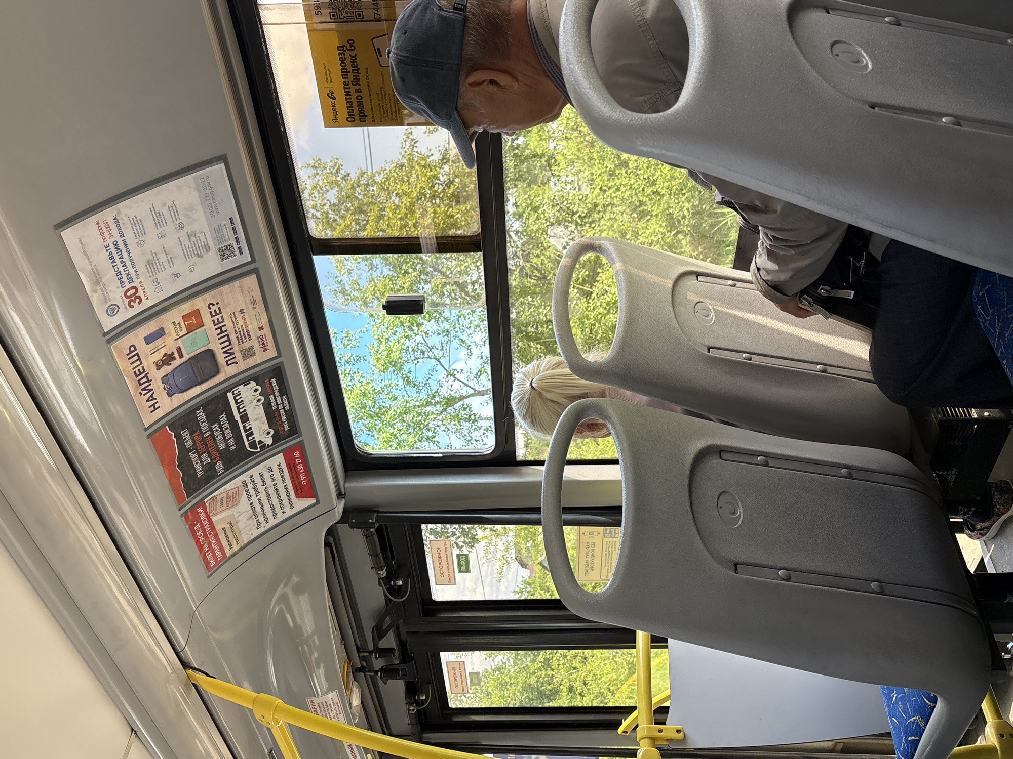 The image depicts the interior of a bus in Pankovka, Russia. Sunlight streams through the windows, illuminating the worn plastic seats and the advertisements affixed to the ceiling. A man wearing a baseball cap and a light-colored jacket is seated, facing away from the camera, looking out the window. A woman with blonde hair pulled into a ponytail is partially visible in the foreground, also seated. The bus appears to be in motion, with green trees and foliage visible outside the windows, suggesting a summer or spring day. Advertising posters are prominently displayed, including one for "Yandex Go" offering payment for travel via QR code. Other posters display information about insurance, taxes, and safety notices. The overall impression is a typical scene on public transportation.