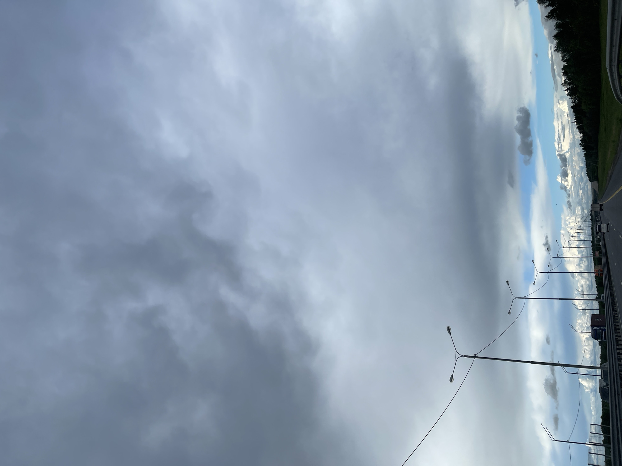 The image depicts an overcast sky dominating the left side, with a highway stretching into the distance on the right. A line of streetlights parallels the road. Trees line the far right edge. Vehicles are visible on the highway. The location is Бавыкино, Russia.