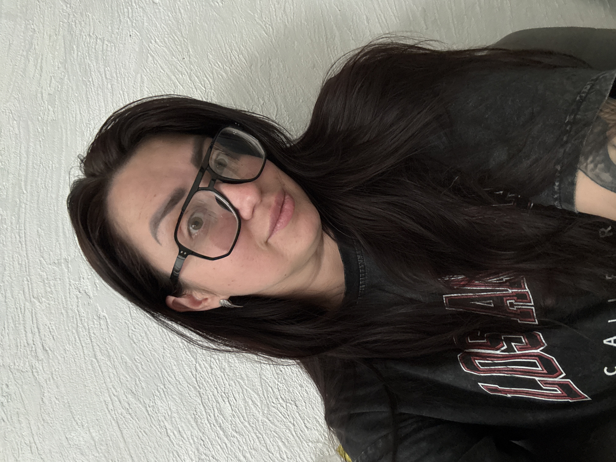 A portrait-style image captures a person, likely a female, from the chest up, looking directly at the camera with a neutral to slightly smiling expression. She has long, dark brown hair that cascades over her shoulders and a dark, possibly grey, patterned t-shirt or sweatshirt. She is wearing black-framed glasses. A small, detailed earring is visible in her left ear (viewer's left).
The person's skin tone is light, and her lips are naturally colored and slightly parted. Her dark eyebrows are visible above her glasses. There's a subtle hint of a tattoo on her right shoulder/arm area, peeking from under her hair and clothing.
The background consists of a textured, light-colored wall, appearing to be white or off-white. The photo is taken at a significant tilt, giving the impression that the subject is reclining or posing dynamically.
Visible text on her dark shirt, in white and red outlined letters, spells out "LOS" and what appears to be "ANGELES" (partially visible as "A" followed by "S") and "CALIFORNIA" (partially visible as "CALI"). The lettering style suggests a collegiate or sports-related theme. The overall setting appears to be indoors, with no specific time of day or weather discernible.