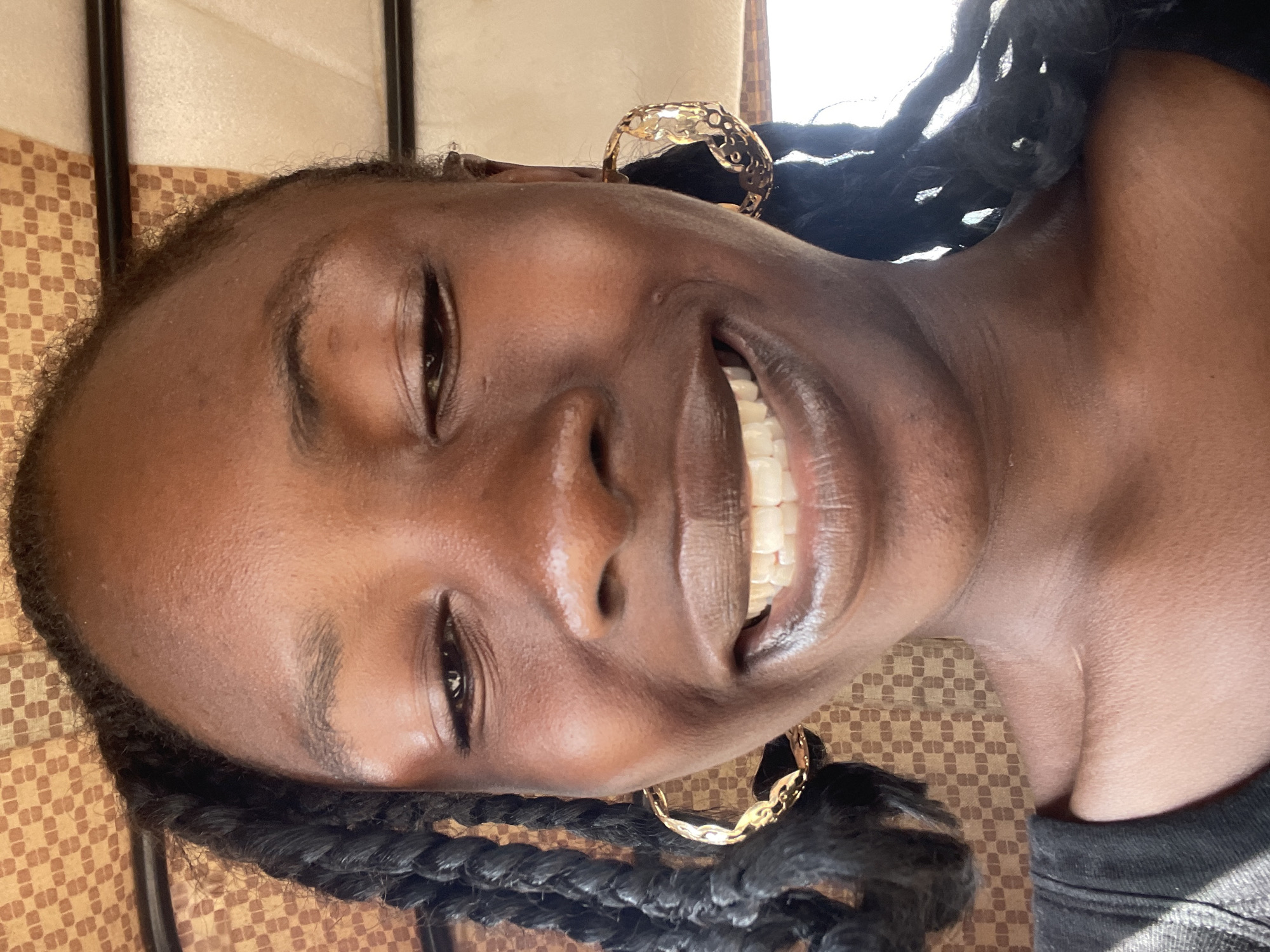 A young Black woman with dark skin and dark hair styled in braids smiles at the camera. She is wearing large, gold hoop earrings. The background is a patterned fabric. The image appears to be a selfie taken indoors.