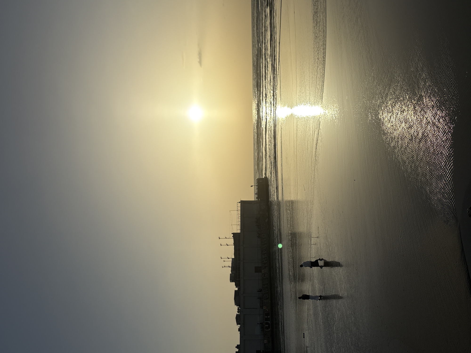 The image captures a beach scene at sunset in an unidentified city. A multi-tiered building stands along the shoreline. Two individuals are walking on the wet sand near the building. The sun is setting over the water, with its reflection visible on the wet sand and in the water.