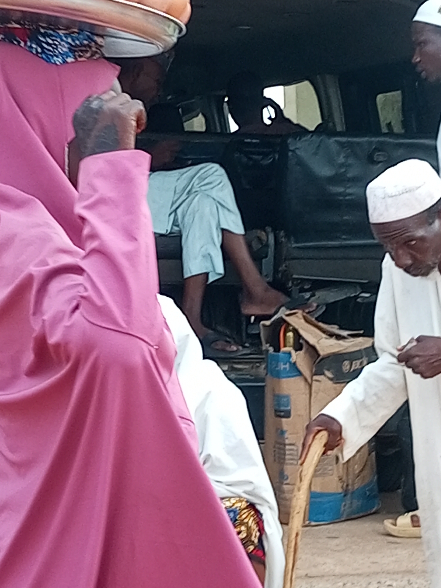 The image depicts a scene in Gombe, Nigeria, with several individuals and objects. In the foreground, a person wearing a vibrant pink garment is partially visible, with a tattoo on their hand. To the right, an elderly man in a white robe and cap holds a wooden walking stick. Behind him, another person in a white robe and cap is also visible. The background reveals the interior of a vehicle, possibly a van, with dark seating. Several large sacks, one appearing to be a cement bag with blue and brown markings, are present in the rear of the vehicle. A gas cylinder is also visible next to the sacks. The ground appears to be a sandy or dirt surface. The overall atmosphere suggests an outdoor setting, possibly a market or a transit point, given the presence of goods and people engaged in some form of movement or waiting.