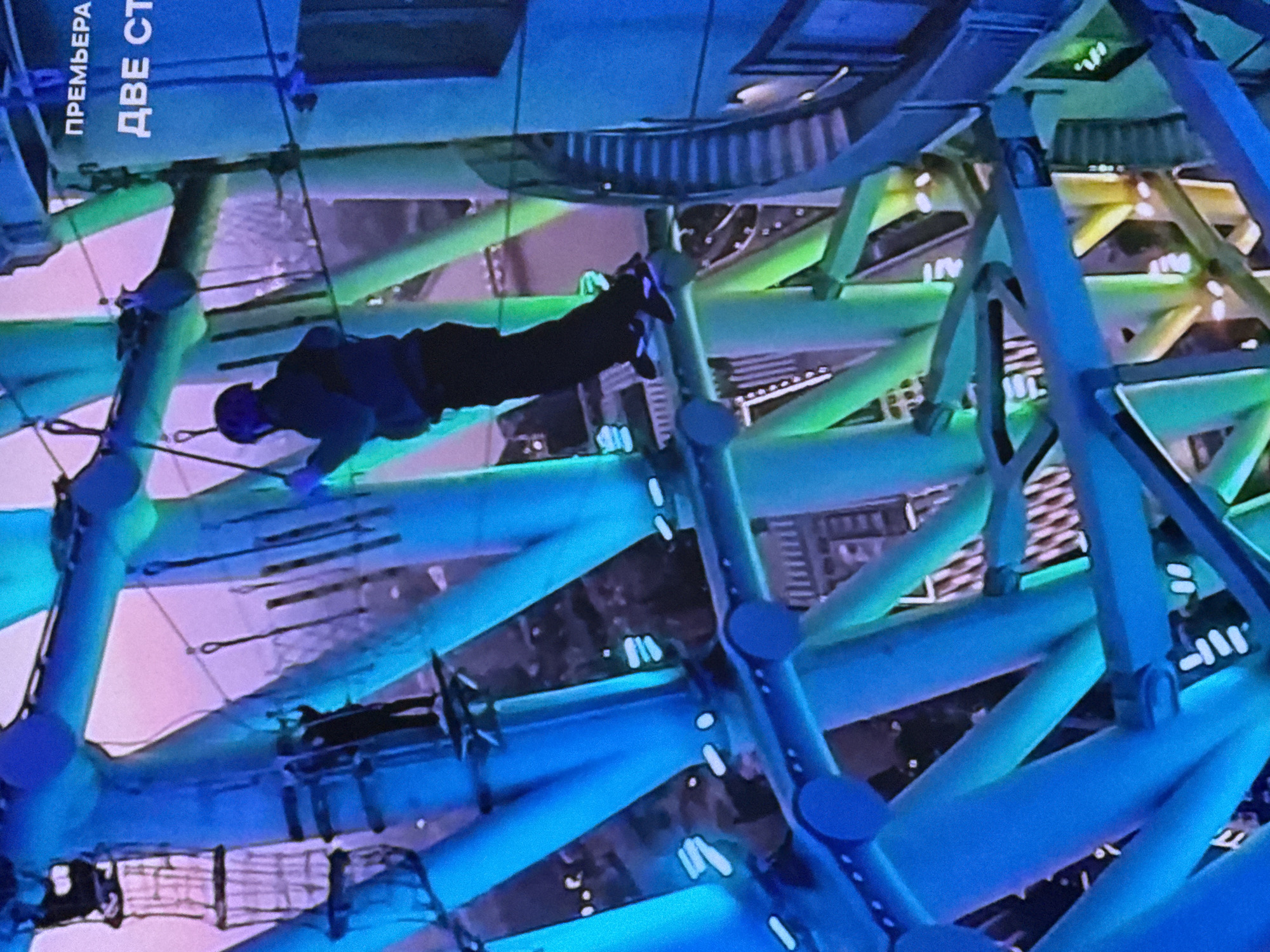 The image shows a person on an elevated walkway, possibly an observation deck or a themed attraction, at night. The structure is made of blue and green illuminated metal beams, creating a vibrant and futuristic atmosphere. In the upper right corner, text in Russian is visible, reading "ПРЕМЬЕРА" and "ДВЕ СТ". This suggests that the location might be related to a premiere event or an attraction with a name that translates to "Two Stairs" or "Two Sides." The background is dark, indicating it is nighttime, and some blurred lights from the city are visible. The person is wearing dark clothing and appears to be harnessed for safety, suggesting they are engaged in an activity like rock climbing or a high-ropes course. The overall scene conveys a sense of adventure and urban exploration in a brightly lit, modern environment.
