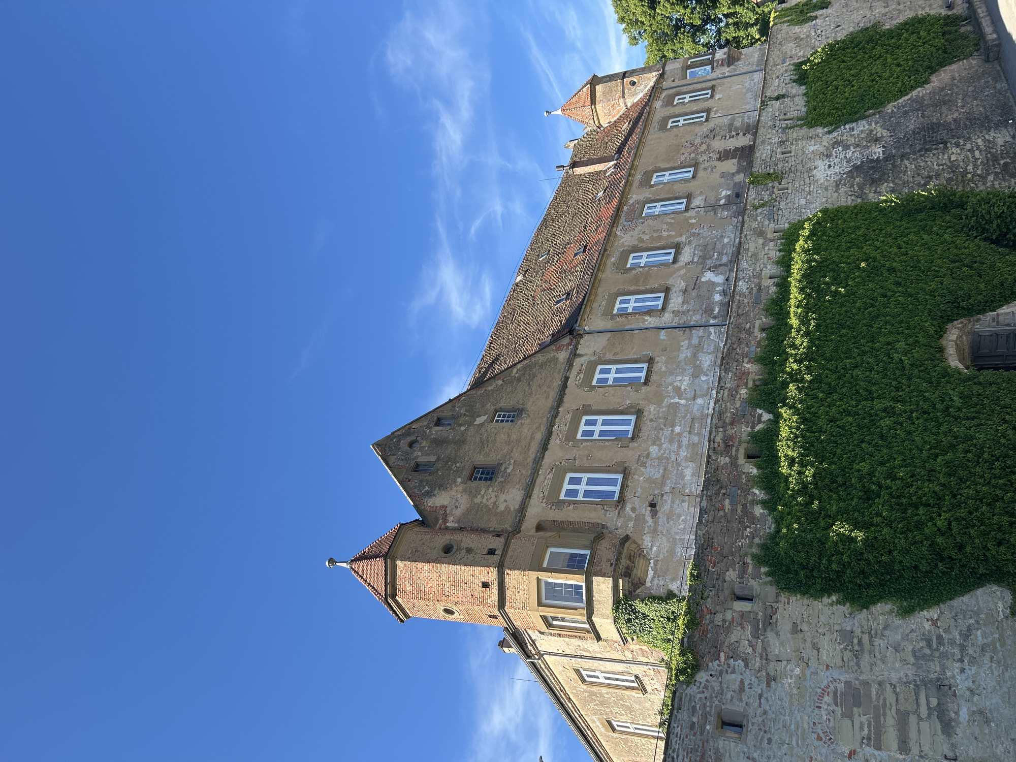 A low-angle perspective shot captures a large, historic building, likely a castle or fortified structure, set against a clear blue sky with wispy clouds. The building's facade features multiple windows, some with white frames, and is partially covered in dense green ivy. The architecture incorporates brick elements, including a prominent tower with a pointed roof. The building's walls show signs of age and wear, with visible brickwork and varied textures. The location is identified as Untergruppenbach, Germany.