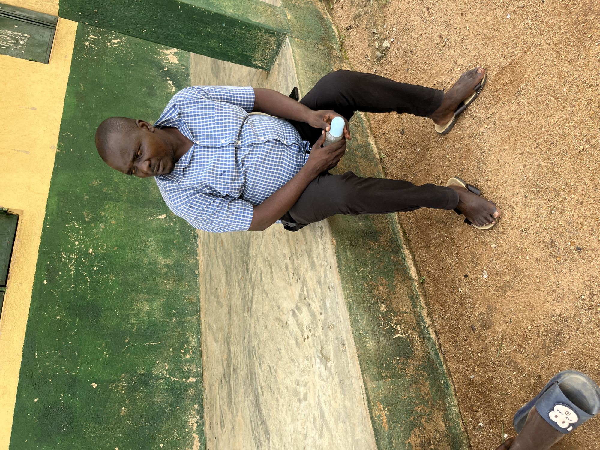 A man is seated outdoors on a raised concrete ledge in front of a textured green and yellow painted wall. He is wearing a blue and white checkered short-sleeved shirt and black trousers. He is looking off to his left. In his hands, he is holding a light blue cylindrical object. He is wearing dark-colored sandals. The ground in front of the ledge is covered in loose sand and small pebbles. Another person's foot wearing a blue sandal is visible on the lower left side of the image. The scene appears to be located in Bukuru, Nigeria.