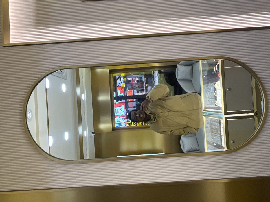 An individual is captured in a mirror selfie within a retail establishment. The subject, a male, appears to be of African descent, wears a tan puffer jacket, and holds a smartphone in his right hand, positioned to photograph his reflection. The mirror is oval-shaped with a gold-colored frame, mounted on a light-colored ribbed wall panel.
The reflection in the mirror reveals the interior of what appears to be a jewelry store, featuring multiple glass display cases exhibiting various items. Two light-colored, armless chairs are also visible in the reflection. The ceiling is white with recessed lighting, and a security camera is mounted near the top of the reflected space.
Beyond the immediate interior, the mirror reflects an outdoor street scene, suggesting the store's entrance or window. This exterior view is illuminated by numerous brightly lit neon signs displaying Chinese characters. A prominent red vertical sign visible in the reflection clearly shows the characters "江津" (Jiangjin), indicating the location is in the city of Jiangjin, China. A light-colored vehicle is partially visible on the street.