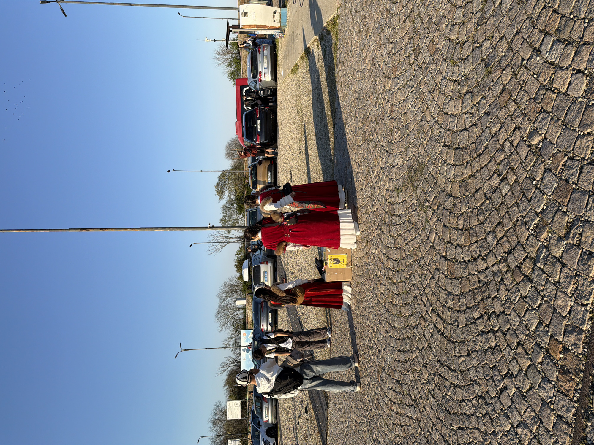 On a cobblestone surface in Unknown, Unknown, three individuals stand next to each other. Two are women wearing traditional clothing with red and white elements. Next to them are two younger people dressed in casual attire. Behind them, cars are parked. In the background are some streetlights and trees under a clear blue sky.