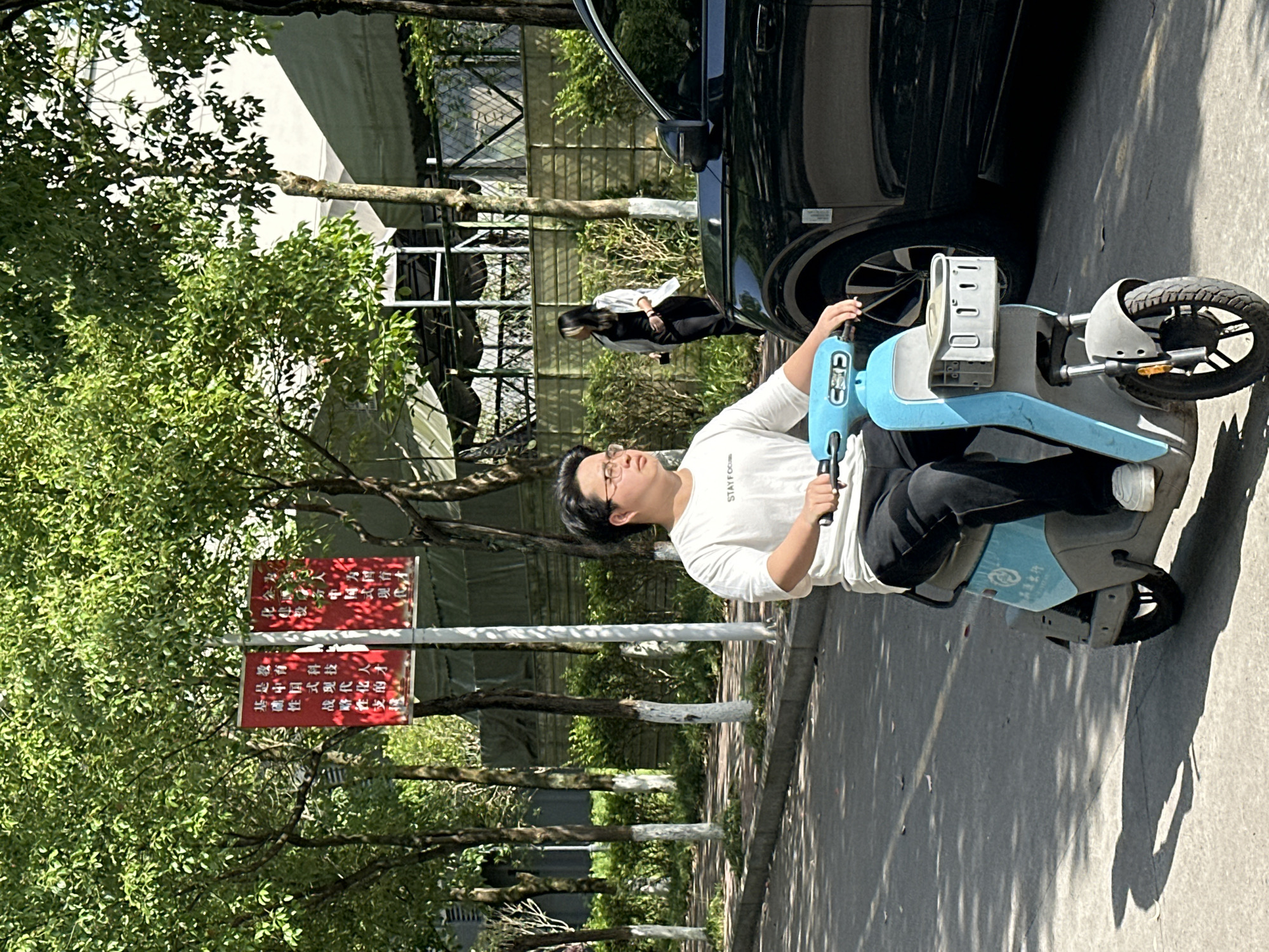 This bright, sunny daytime image captures an outdoor street or pathway scene, likely in Jiangjin, China. The primary subject is a man sitting on a light blue and gray electric scooter, holding its handlebars and looking upwards. He wears a white short-sleeved t-shirt with the partial text "STAY FOCU" visible, and dark pants, along with eyeglasses.
In the background, a black car is parked, and another person, appearing to be a woman with long dark hair, is standing near the vehicle, dressed in a white top and dark pants. The setting is characterized by numerous green trees and lush foliage, casting strong shadows on the paved ground, indicative of clear weather.
On the left side of the frame, two red rectangular signs are mounted on a pole amidst the trees. The Chinese text on these signs translates to themes such as "Chinese-style modernization," specifically mentioning "education, science and technology" as its "foundational, strategic support." The overall scene suggests a quiet moment, with the man seemingly resting or observing something above him.