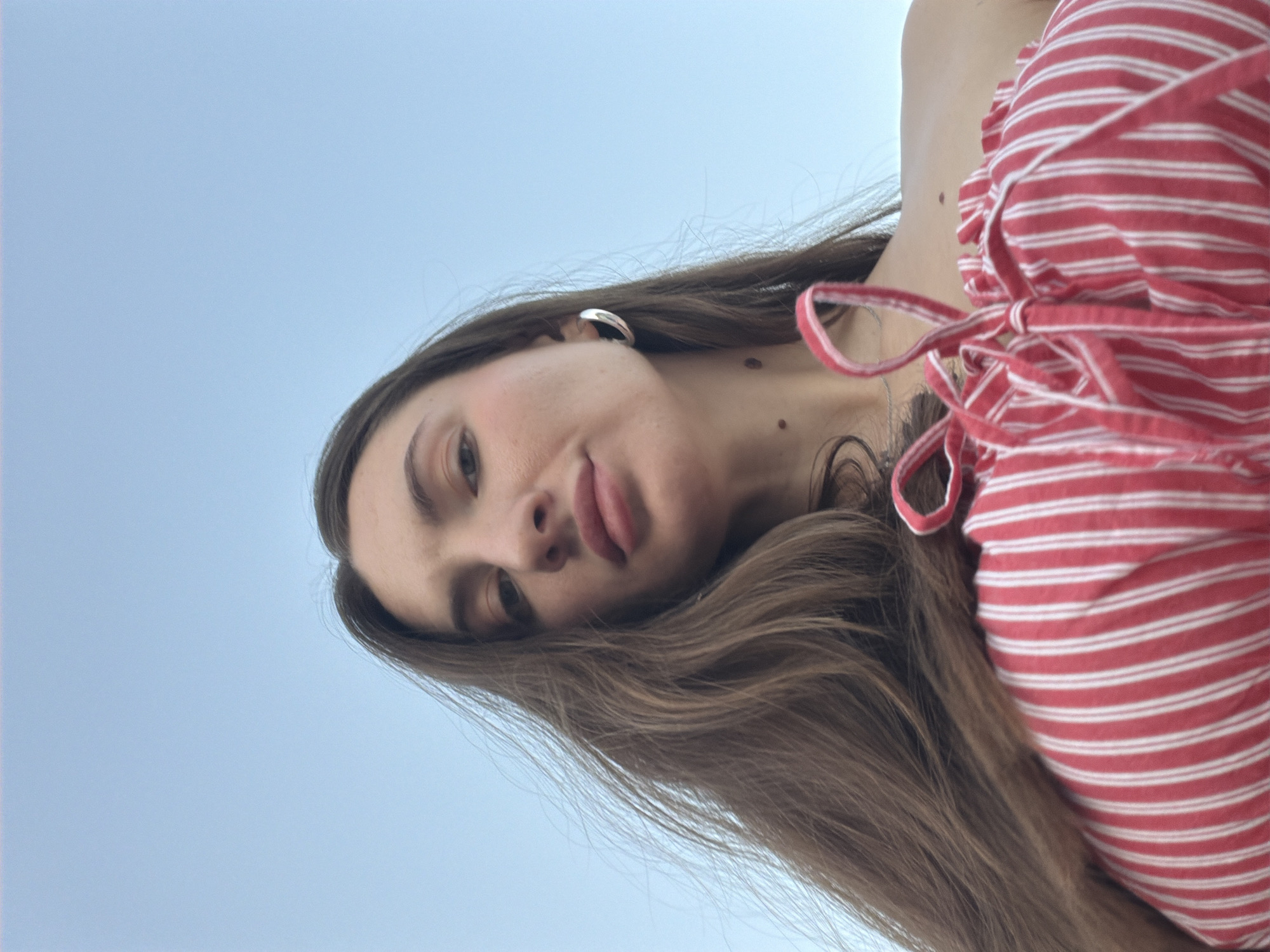 A low-angle, close-up, selfie-style shot captures a young woman against a clear, pale blue sky, likely taken outdoors in Sharm El Sheikh, Egypt. The woman has long, light brown hair that is blowing in the wind, framing her face. She is wearing a red and white striped top with thin straps tied at the front, revealing her shoulders. A large, silver hoop earring is visible on her left ear. Her facial expression is neutral, with her lips slightly parted. The background is a vast expanse of sky, indicating an open area with no discernible city structures.