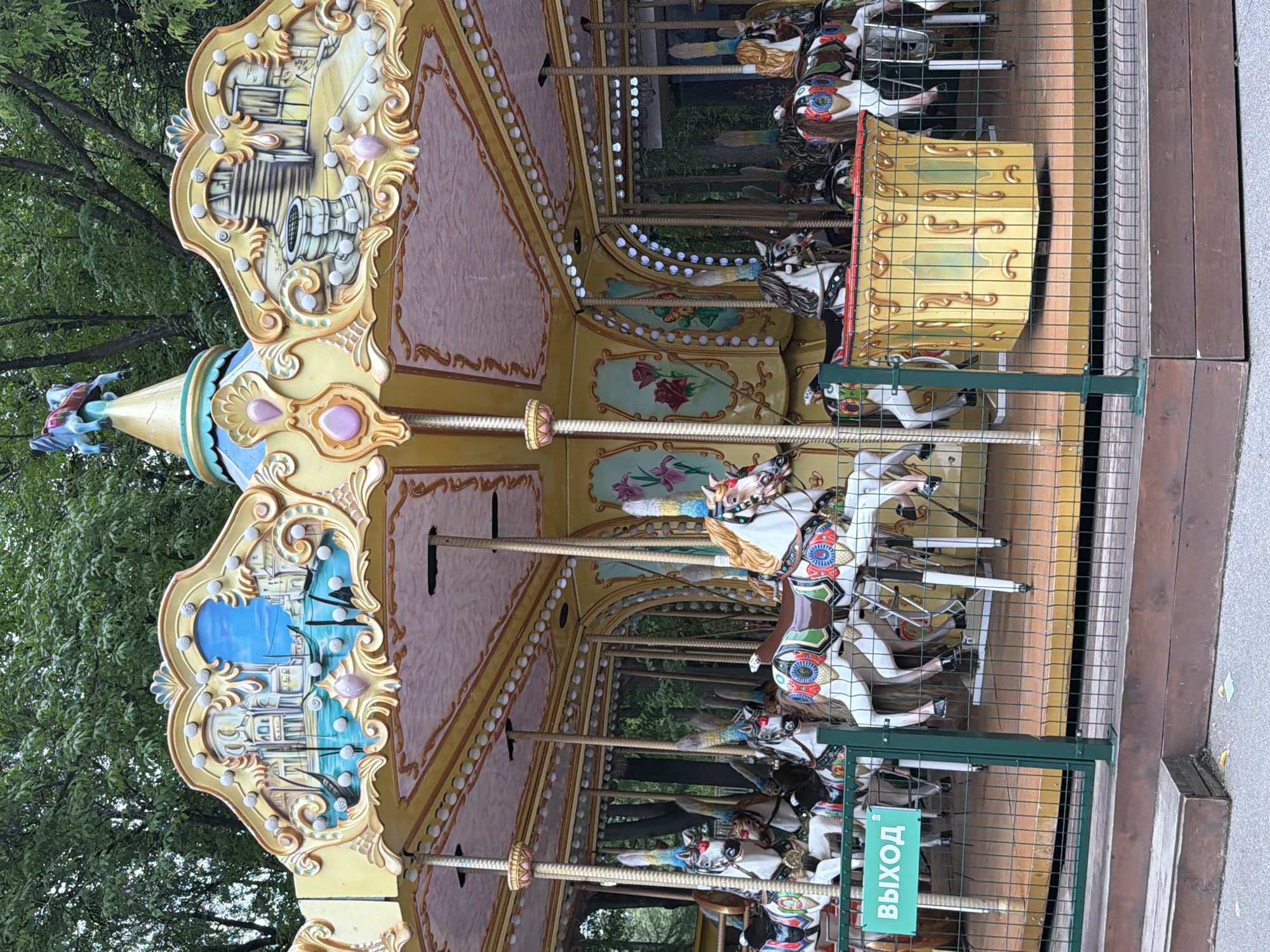 This image shows a brightly decorated carousel with multiple horses. The carousel is set within a park or outdoor recreational area, with trees visible in the background. A green sign with the word "ВЫХОД" (Exit in Russian) is attached to a metal fence in the foreground. The carousel itself features intricate artwork on its upper panels, depicting scenes that appear to be of a city and a well. The horses are ornately decorated, each with unique saddle and bridle details. The overall scene suggests a public attraction, likely a family-friendly entertainment venue. The location is the Perm City Park (Gorodskoy Sad) in Perm, Russia.