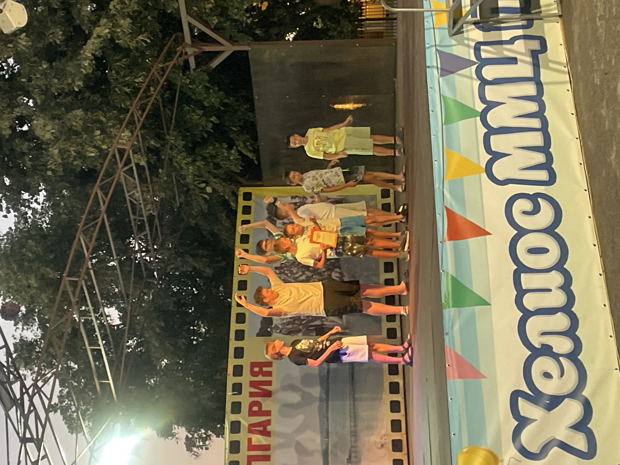 A group of young boys stand on a stage in front of a backdrop with the word "BULGARIA" written on it in large red letters. The boys appear to be participating in an event, with some holding certificates or awards. They are dressed in casual summer clothing, and most of them are smiling. The stage is decorated with colorful banners. In the background, trees are visible, suggesting an outdoor setting. The overall atmosphere is festive and celebratory. The event appears to be taking place in Kiten, Bulgaria.