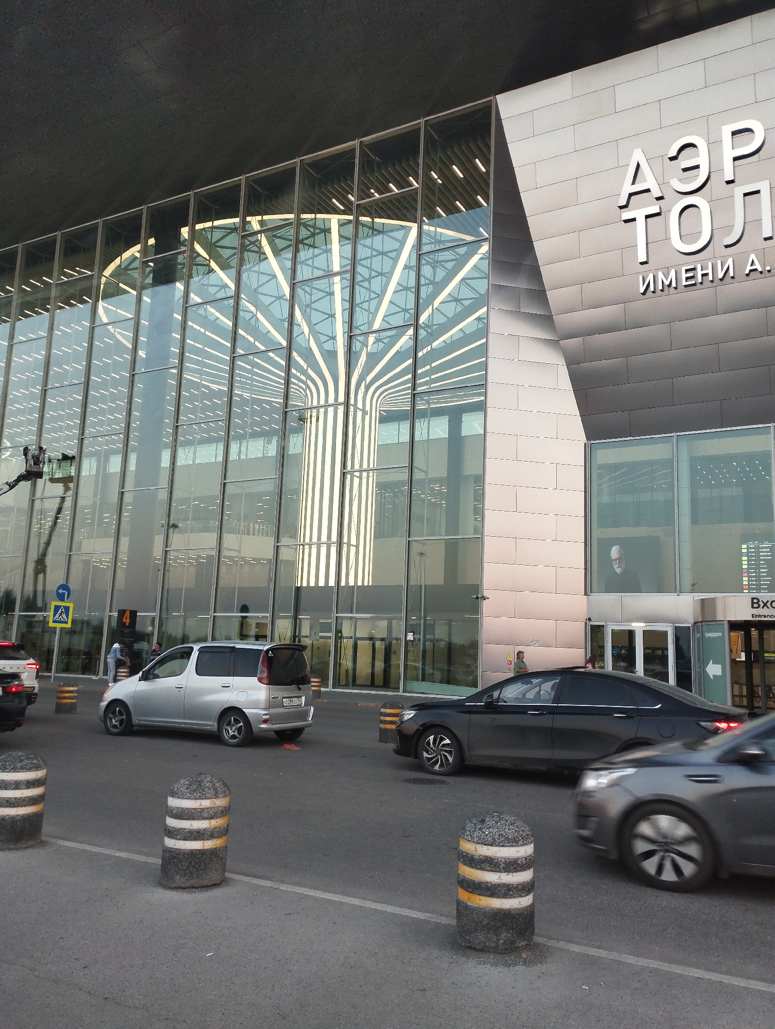 The image shows the exterior of Tolmachevo Airport in Ob, Russia. The airport building features a modern design with large glass windows and metallic facade panels. The text "АЭРО" and "ТОЛ" with "ИМЕНИ А." below it, signifying "TOLMachevo", is visible on the building's facade. Several cars are on the road in front of the terminal, including a silver minivan, a black sedan, and a partially visible white car. Bollards with white and yellow stripes are placed along the road. Inside the terminal, seen through the glass, there is an illuminated tree-like structure. A large screen displaying a portrait of a man with a beard is visible on the right side of the building. A person appears to be working on the exterior of the glass facade on a lift. Several people are visible near the entrance of the building.