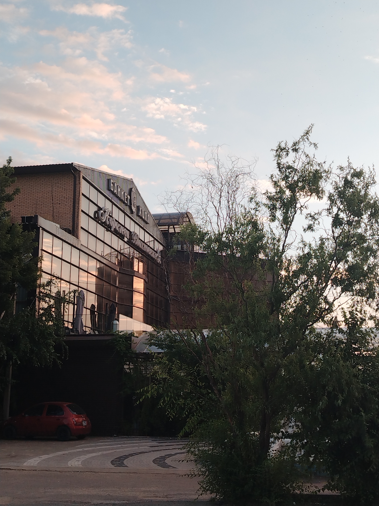 The image captures a building exterior in Krasnodar, Russia, with signage that appears to read "Банкет Холл" and "Курортный". The building features a facade of reflective glass and brick. In the foreground, a red compact car is parked on a patterned driveway. Lush green trees partially obscure the view of the building and driveway, with a cloudy sky overhead.