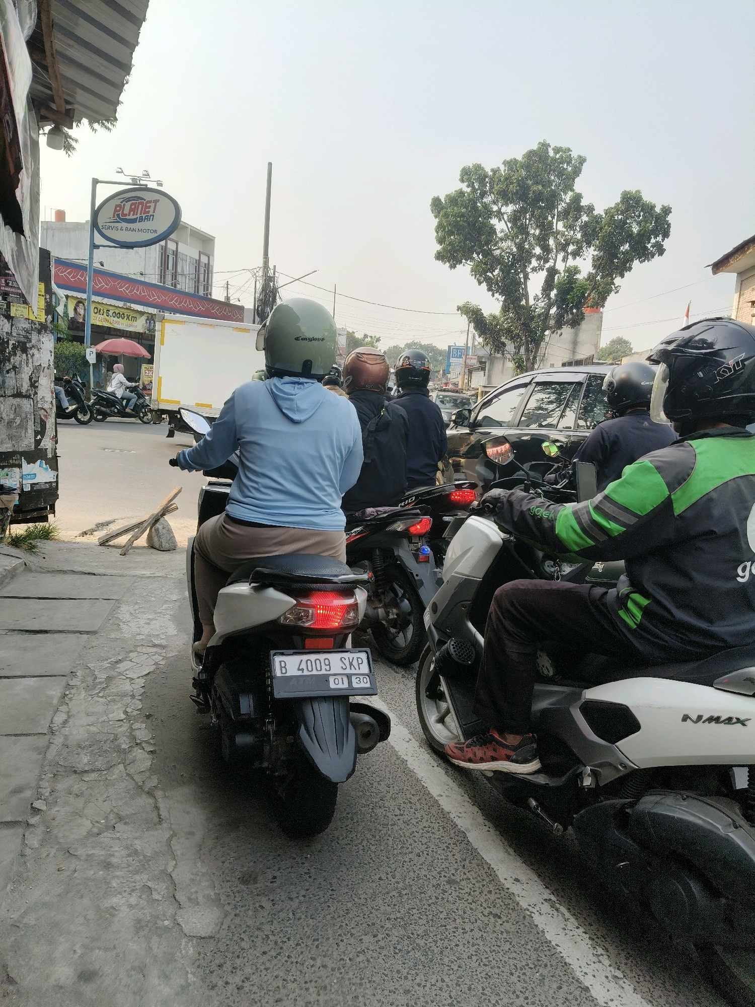 The image captures a street scene in South Tangerang, Indonesia, likely during daylight hours, as suggested by the bright sky. Several individuals on motorcycles are waiting in traffic, creating a typical urban congestion scenario. In the foreground, a person wearing a light blue long-sleeved shirt and brown pants is seated on a white scooter. The scooter's license plate is visible, reading "B 4009 SKP" with smaller text "01 30" below it. To the right, another individual is on a white scooter, wearing a black and green jacket with reflective stripes, indicative of a delivery service uniform. This person has on red and black patterned sneakers. Behind them, other motorcyclists are present, one wearing a brown helmet and another a black helmet. In the background, a black car is stopped in traffic. A large green tree stands prominently in the mid-ground. Signs are visible on the left side of the image, including one that says "PLANET BAN SERVIS & BAN MOTOR". A white truck with "OU: 5.000 KM" written on it is also present. The environment appears to be a street with a sidewalk and building structures on the left, and the road with traffic on the right. The overall atmosphere suggests a moment of pause in daily urban transit.
