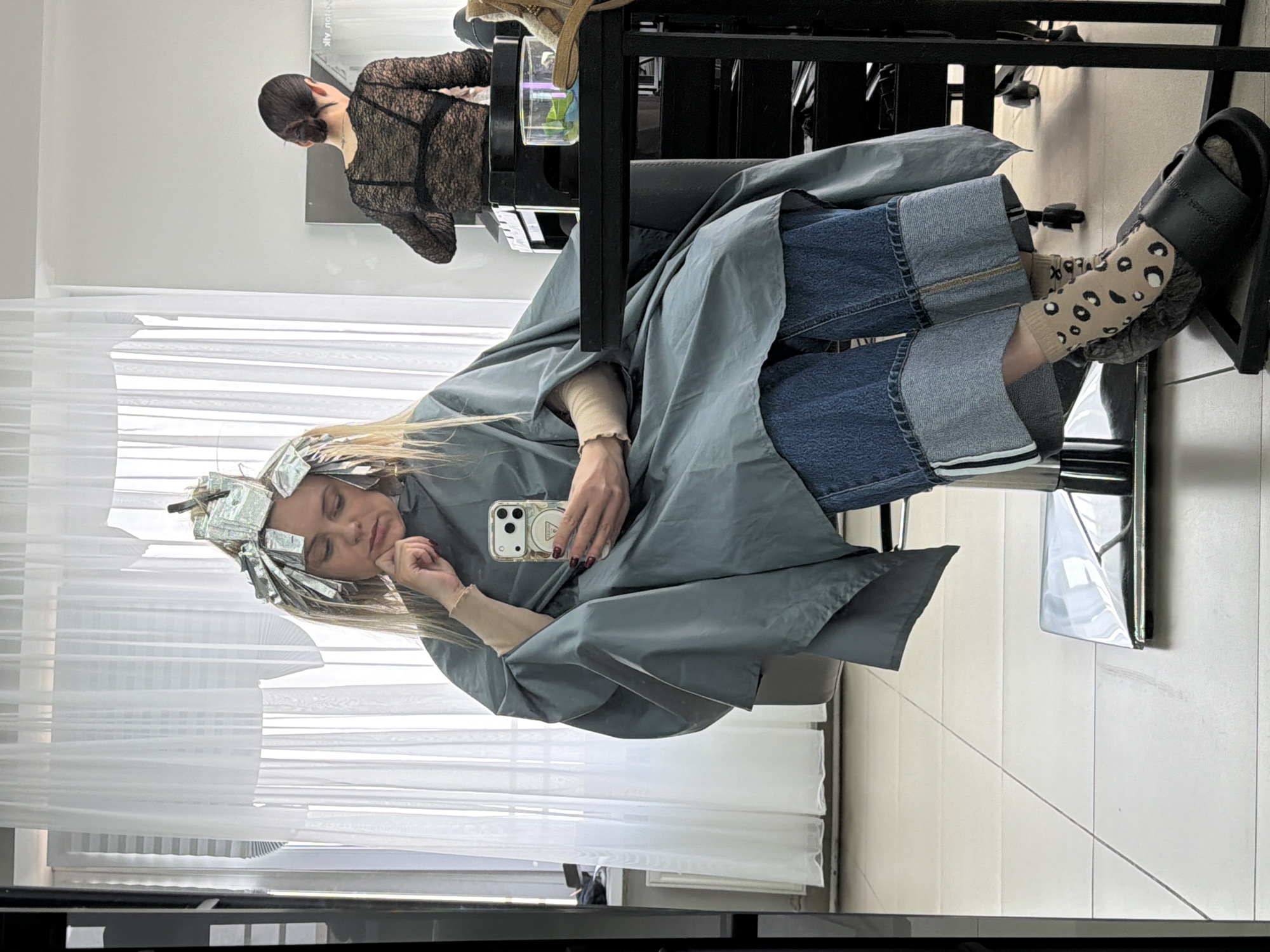 The image shows a woman with foil in her hair, seemingly undergoing a hair coloring treatment at a salon. She is sitting in a chair, draped in a gray cape, and holding a smartphone. She appears to be resting her head on her hand, perhaps waiting for the hair dye to process. Her outfit consists of blue jeans with cuffed hems, patterned socks, and black sandals. In the background, another person wearing a black lace top is visible, likely a hairdresser working in the salon. The salon appears to have white walls and a large window with sheer curtains. The location is identified as the city of Votkinsk, Russia, but there are no specific landmarks visible in the image to confirm the exact location within the city.