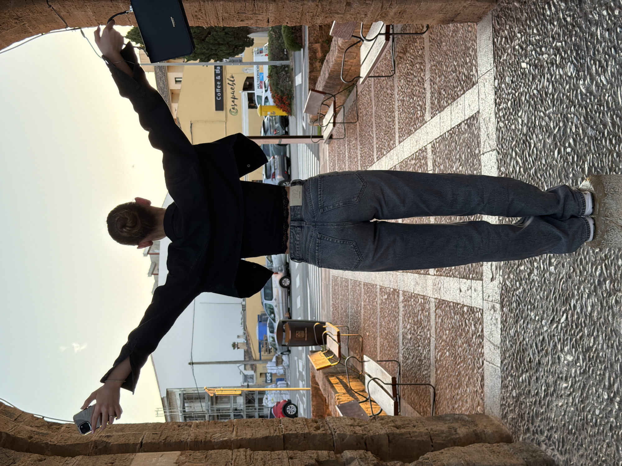 A person stands in a stone archway, arms outstretched, holding a black purse in their left hand and a cell phone in their right. The person is wearing a black jacket and gray jeans. The archway opens onto a street in Alcúdia, Spain, with benches lining the walkway and buildings visible in the background, including a business sign for "Coffee & Bla...". The ground is paved with a textured stone pattern. Cars and crosswalks are visible in the street.