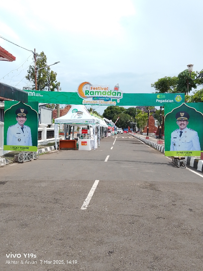 The image shows a street in Indramayu, Indonesia, located at latitude -6.326, longitude 108.323. The street is lined with white painted curbs and black and white striped markings. A banner archway spans the street, decorated with a large, green banner that reads "Festival Ramadan" in large, white lettering. The banner has a smaller, green banner underneath that reads "Ramadan Gemilang" in white lettering. The archway is decorated with large, green posters with the images of men in uniforms on either side. The posters on the left are titled "LUCKY HAKIM" and on the right "SYAEFUDIN." Between the posters, numerous white tents line the street. The tents have been set up for a Ramadan Festival. A few people can be seen walking through the festival. The sky is clear and blue. A white sedan is driving away from the camera, and a second, white sedan is parked further down the street.