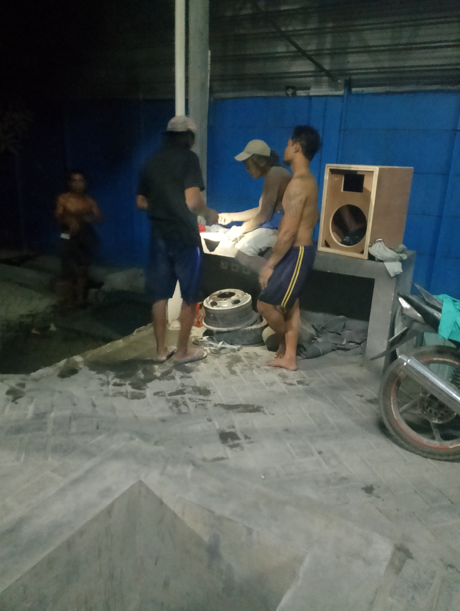 The image depicts an outdoor scene at night, possibly in Jambean, Indonesia, with three men present. One man, wearing a dark t-shirt and shorts, stands facing right, holding an object. Another man, shirtless and wearing shorts, stands facing left. A third man, also shirtless and wearing shorts, is seated facing right. In the foreground, a concrete floor is visible with some water stains. In the background, there is a blue wall and a structure resembling a table with a wooden speaker box on top. A motorcycle is partially visible on the right side of the image. The environment appears to be a workshop or an outdoor storage area.