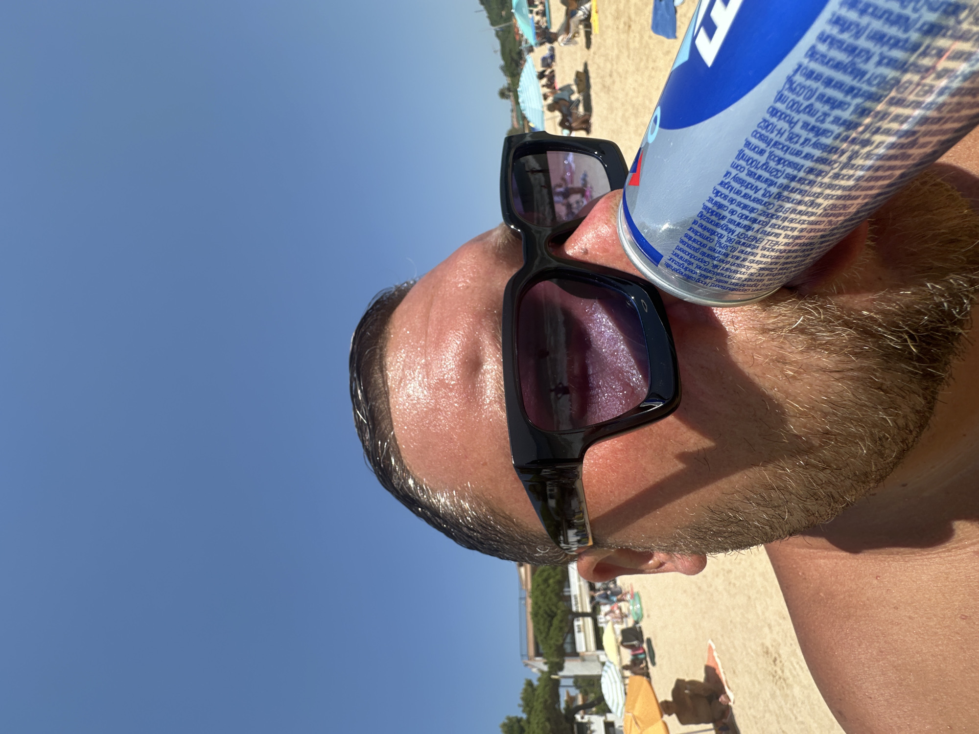 A close-up, eye-level, outdoor shot shows a person wearing black, rectangular sunglasses and a light beard, drinking from a blue and silver can. The person's head and shoulder are visible, with their skin appearing tanned. In the background, a sandy beach with beach umbrellas, lounge chairs, and people is visible under a clear blue sky. The location appears to be Sant Feliu de Guíxols, Spain, as indicated by the visible buildings and beach scene characteristic of the Costa Brava. The lighting suggests it is a sunny day.