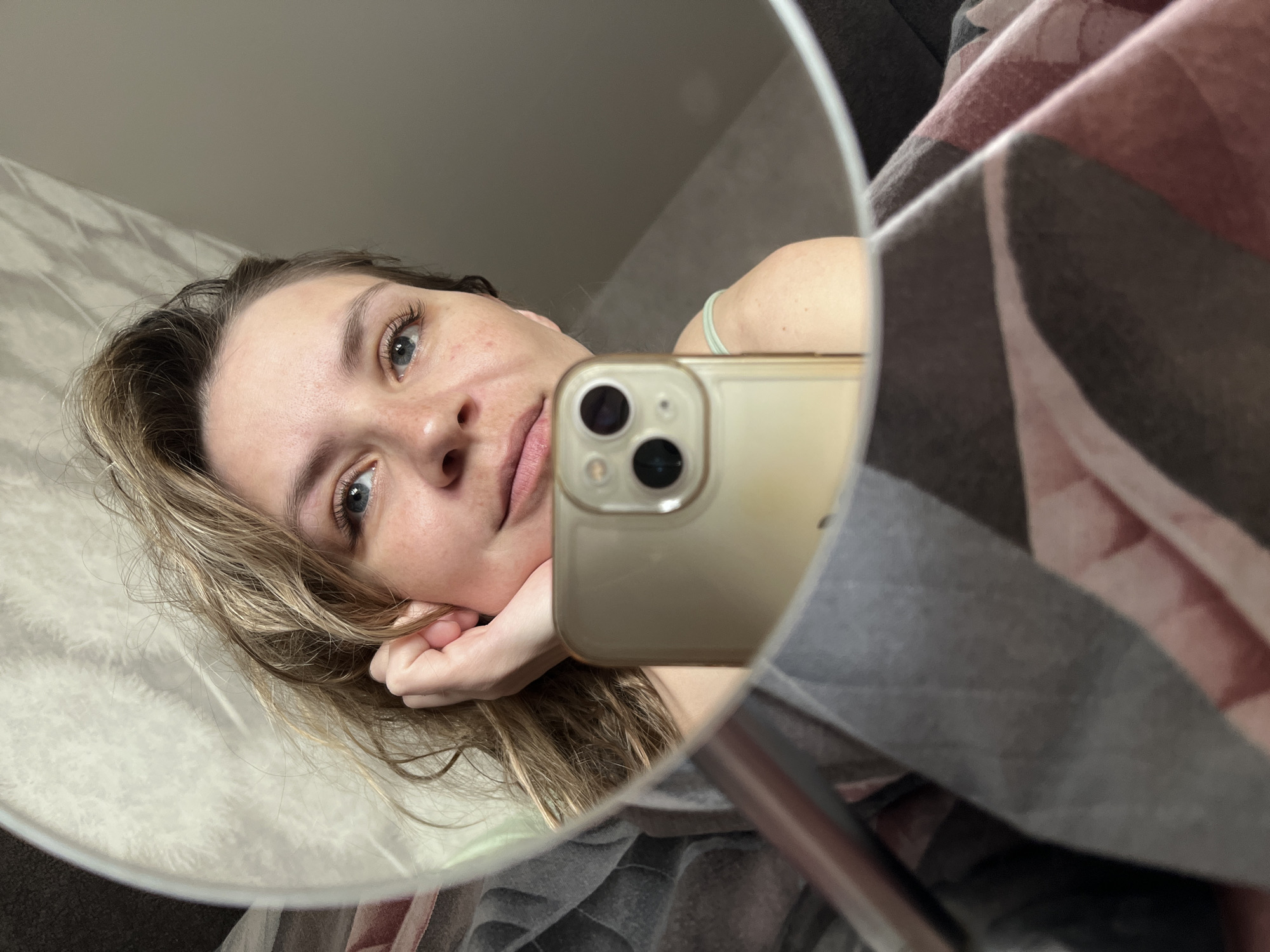 An adult female is depicted in a mirror selfie, reflecting an indoor setting in Chaikovsky, Russia. She has light skin, wavy light brown hair, and blue eyes, with minor freckling visible on her nose and cheeks. Her right hand supports her chin. She is holding a gold-colored smartphone, encased in clear material and featuring a triple-lens camera array, positioned in the lower-center foreground, which is capturing the reflection. A patterned blanket or duvet in shades of dark gray, light gray, and light pink/maroon with geometric shapes partially covers the lower portion of the frame. A light-colored (possibly mint green) strap or garment is visible on her right shoulder. The reflected background consists of a light-colored wall or curtain with a subtle, irregular texture.