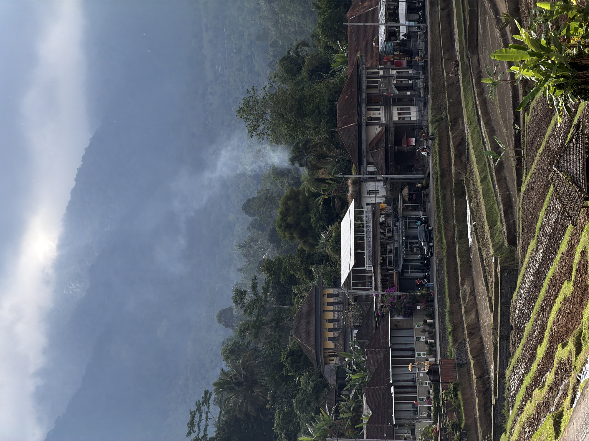 The image shows a view of the village of Penebel, Indonesia, nestled between lush green rice terraces and a misty, forested mountain range. In the foreground, tiered rice paddies are visible, some with water, others recently tilled, and some with young green shoots. A few banana trees and thatched-roof structures are scattered amongst the terraces.
In the midground, a cluster of buildings forms the village. These structures have a mixture of traditional and modern architecture, with tiled roofs and some featuring balconies and open-air sections. A road runs through the village, with a few parked cars and motorcycles. People are visible on the road and near the buildings, engaged in various activities like walking and riding motorcycles. Smoke rises from a patch of the forest in the distance, indicating some form of outdoor activity.
The background is dominated by a large, mist-shrouded mountain covered in dense vegetation. The sky above is partly cloudy, with shafts of sunlight breaking through, creating a diffused, atmospheric light. The overall impression is of a tranquil, rural setting in a tropical environment.