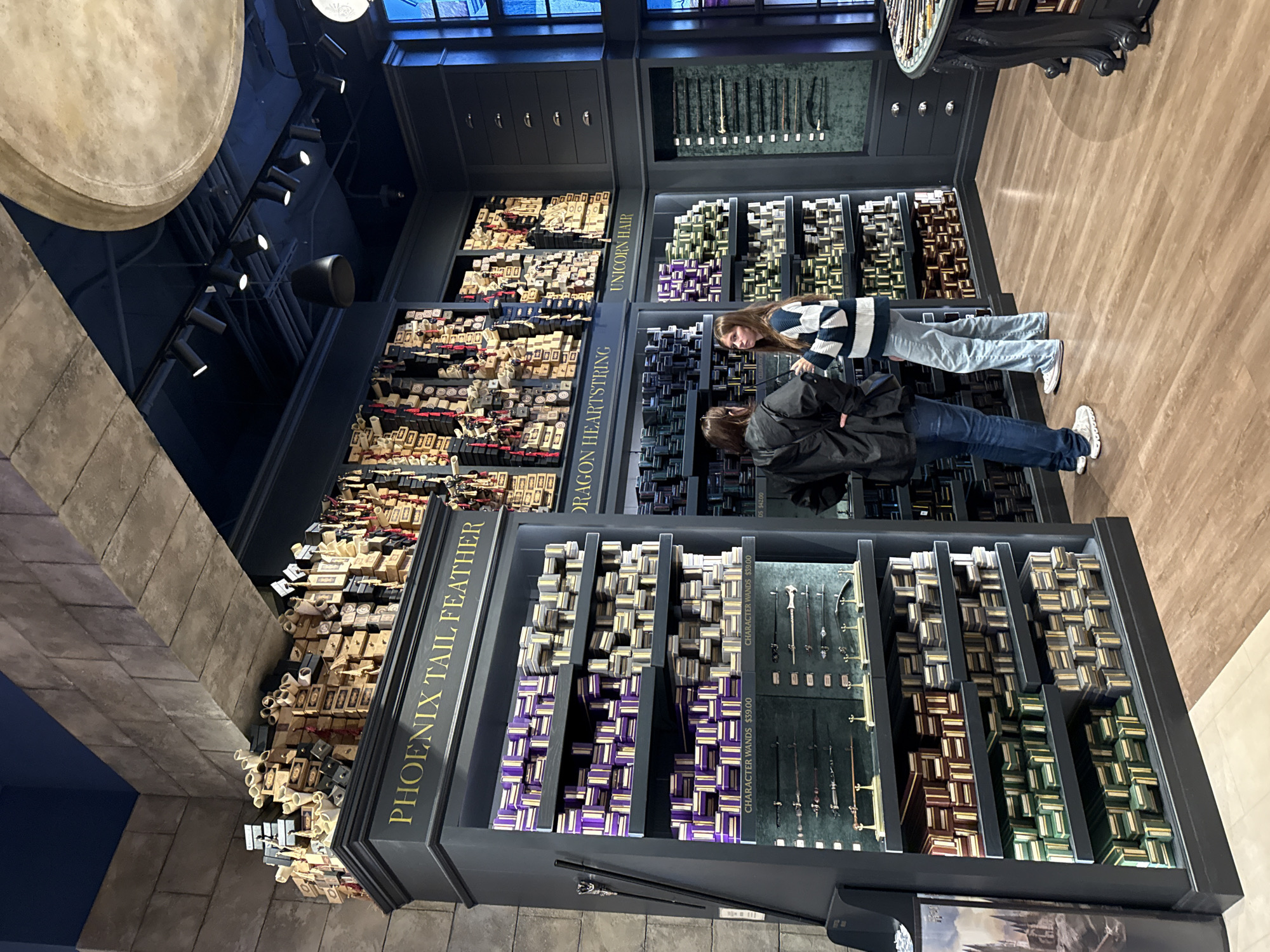 In a well-lit store, two people are browsing a vast collection of wands displayed in dark shelving units. The shelves are organized into sections, each labeled with a name, such as "PHOENIX TAIL FEATHER," "DRAGON HEARTSTRING," and "UNICORN HAIR." Within these sections, numerous wands are neatly arranged in their individual boxes, creating a visually striking display. Some sections feature wands of different colors and designs, while others show specific wands mounted for closer inspection, with prices like "$39.00" and "$42.00" visible.
One person, wearing a blue and white striped sweater and jeans, is leaning in to examine the wands. The other person, in a black jacket and jeans, is standing slightly behind and to the side, also looking at the merchandise. The overall setting appears to be a themed retail environment, possibly related to fantasy or a movie franchise, given the names of the wand sections. The lighting is bright and focused on the displays, highlighting the products. The floor is a polished wooden surface. The image captures a moment of shopping and exploration within this specialized store.