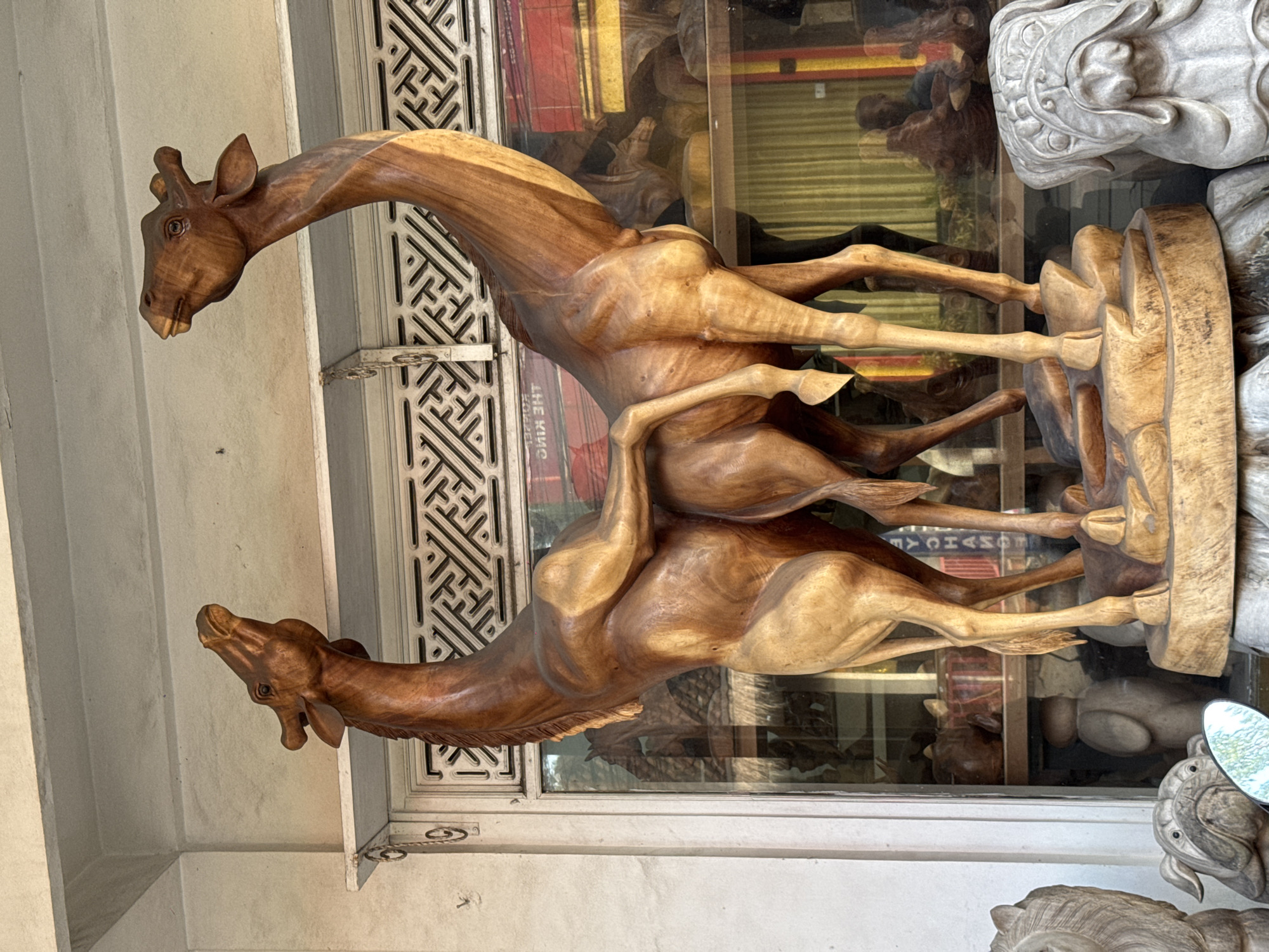 This image depicts a carved wooden sculpture of two giraffes in what appears to be a shop or display area, likely located in the city of Ubud District, Indonesia. The giraffes are carved from light-colored wood and are positioned in a dynamic pose, with their necks intertwined. The sculpture is mounted on a rough, uneven wooden base.
In the background, through a glass window, shelves are visible displaying other carved wooden objects and possibly some fabric items. Reflections on the glass show signage, including text that appears to be in English, and the blurred outlines of other artworks and a person. To the left and right foreground, other carved wooden sculptures are partially visible, including what appears to be a bulldog or similar dog breed. The overall environment suggests a place that sells handcrafted art or souvenirs. The lighting is natural, casting shadows and highlighting the grain of the wood.