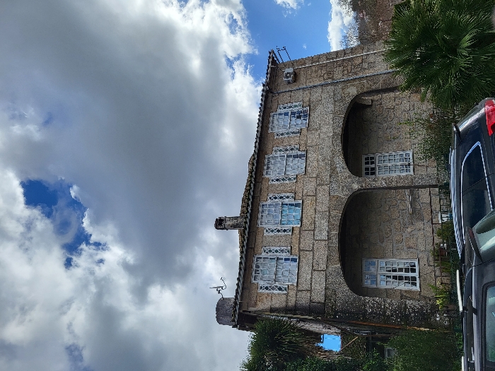 The image displays a two-story traditional Portuguese stone building situated in São Pedro do Sul, Portugal, beneath a partly cloudy sky with patches of blue. The facade is constructed from large, irregular cut stone blocks. The ground floor features two recessed arches, each containing a white-framed, multi-paned window. The upper floor presents five visible white-framed, multi-paned windows, each fitted with a white, decorative metal grille and internal sheer curtains. The tiled roof includes a central stone chimney and, on the left, a small circular stone structure with an antenna. An external air conditioning unit is mounted on the upper right wall. Dense green foliage, including a large palm on the left and a bushy palm in the right foreground, surrounds the building. Two dark-colored vehicles are partially visible, parked in the bottom foreground. No people are present.